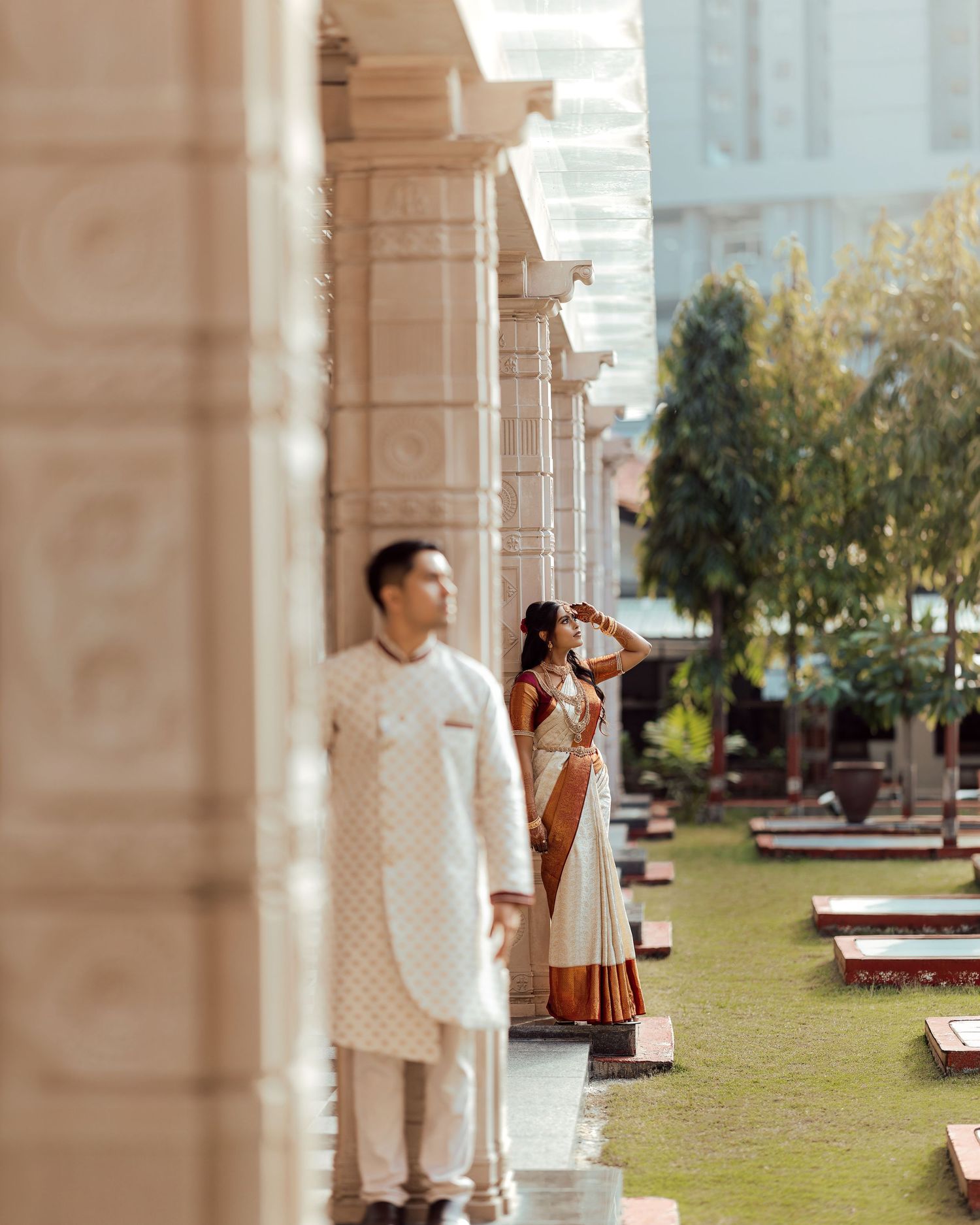 Indian couple in traditional wedding attire posing between ornate temple columns in a lush garden setting.