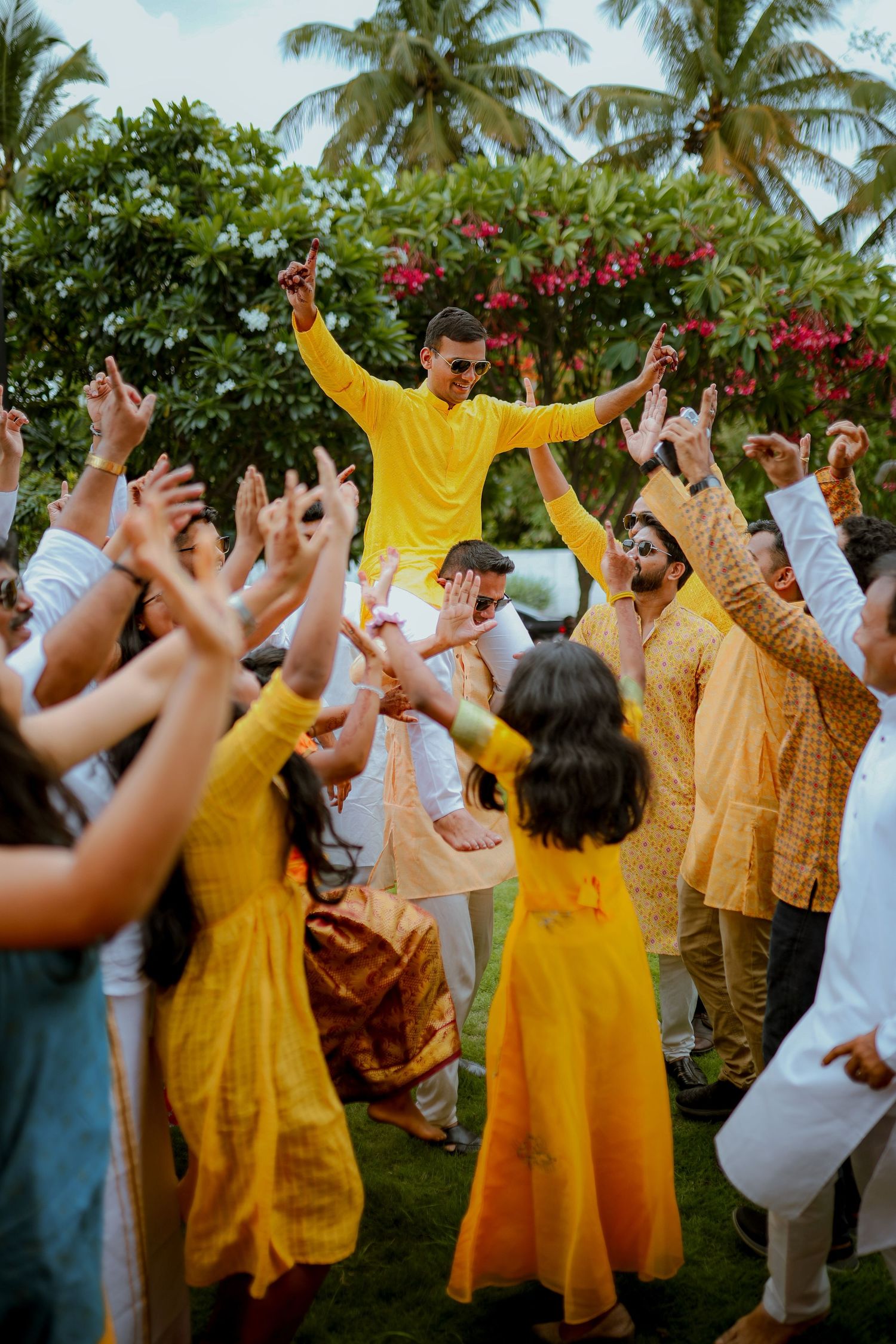 A group of wedding guests in yellow and white attire dancing joyfully at an outdoor celebration with palm trees.