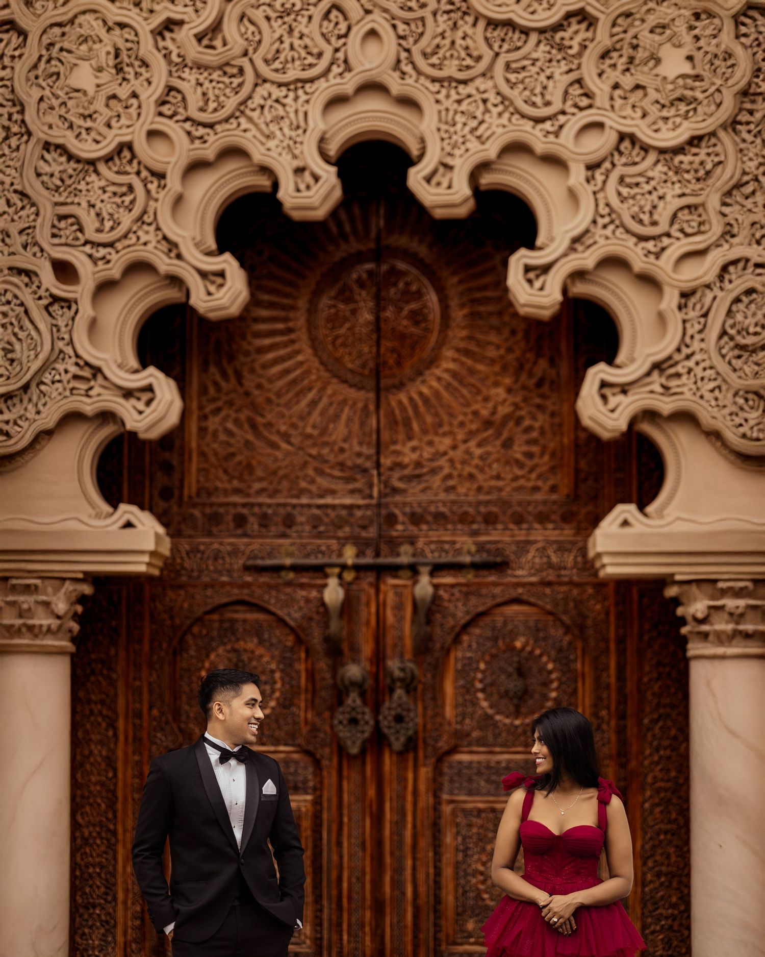 A couple shares a romantic moment in front of an ornate wooden door with intricate architectural details and arches.
