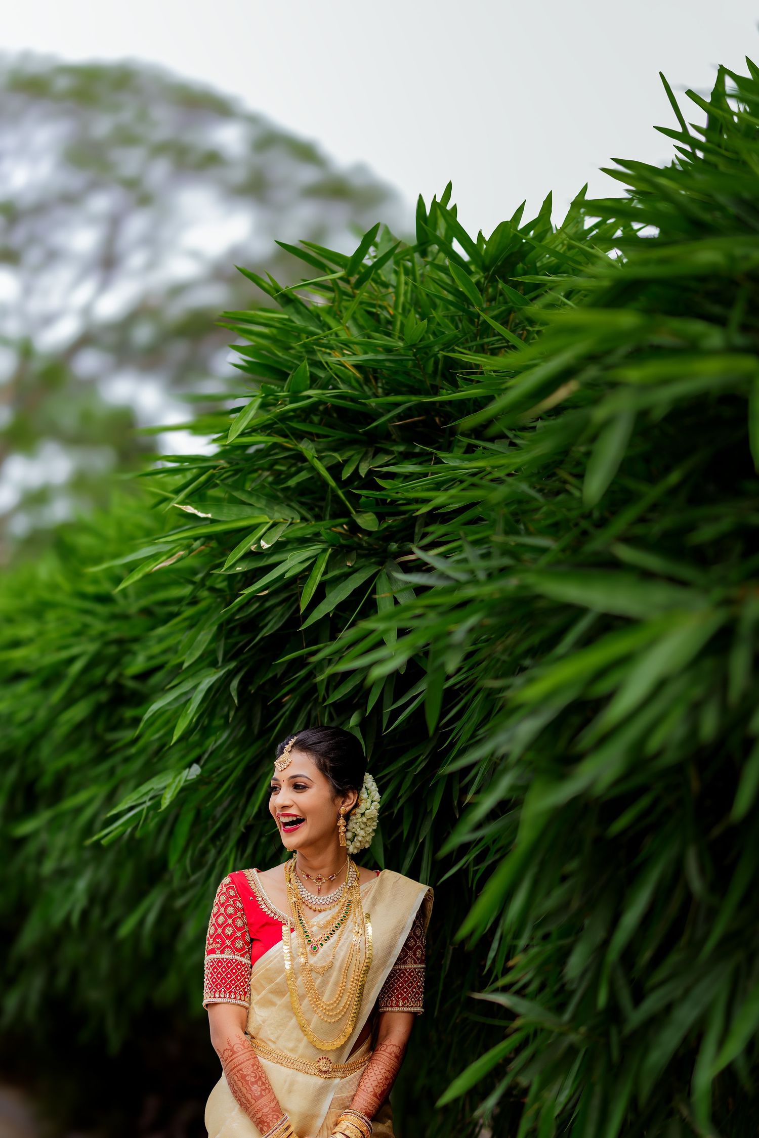 Traditional South Indian bride in cream silk saree and red blouse stands against lush bamboo foliage background.