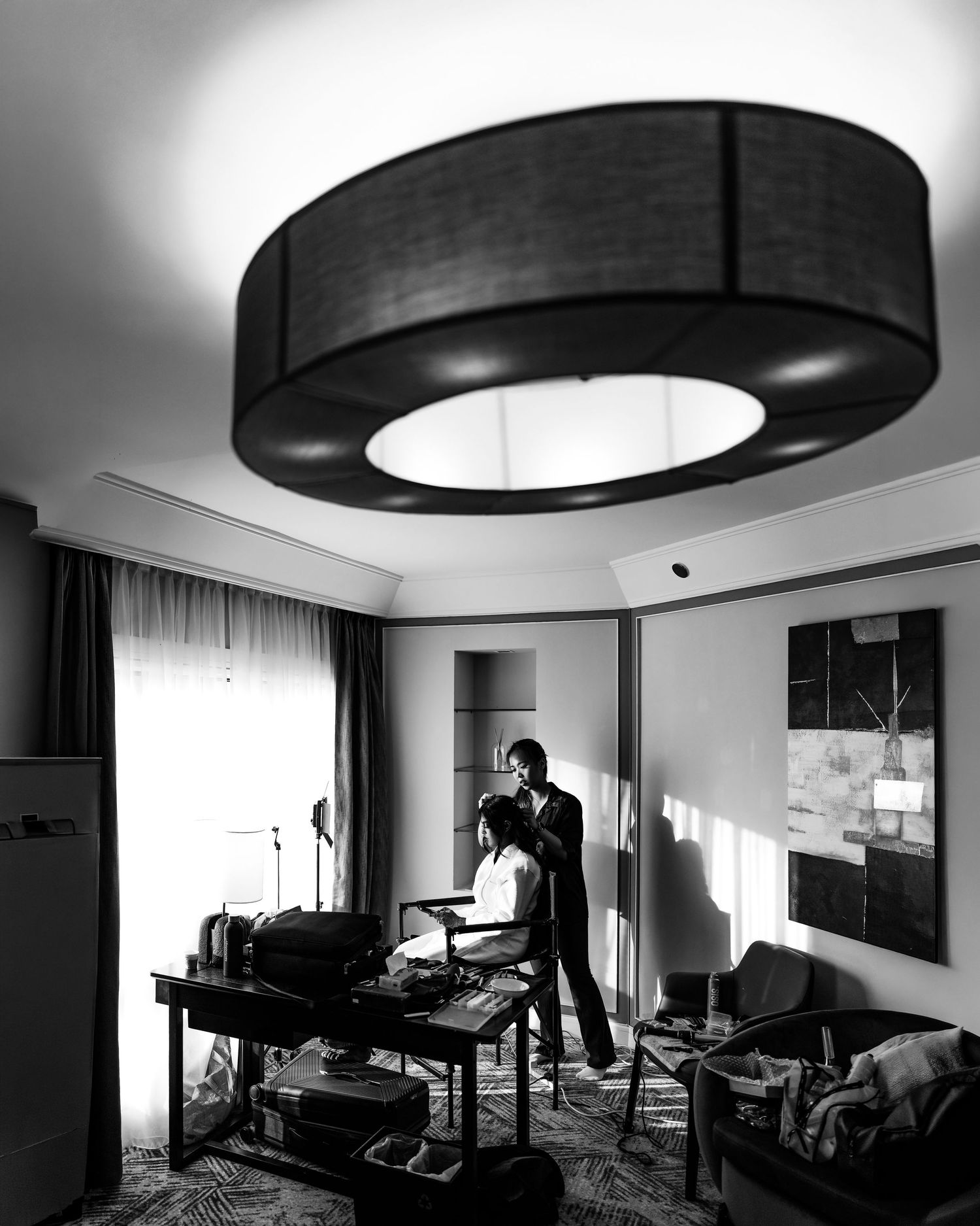 Black and white photo of a stylist working on a seated client in an elegant hotel room with a large drum ceiling light.