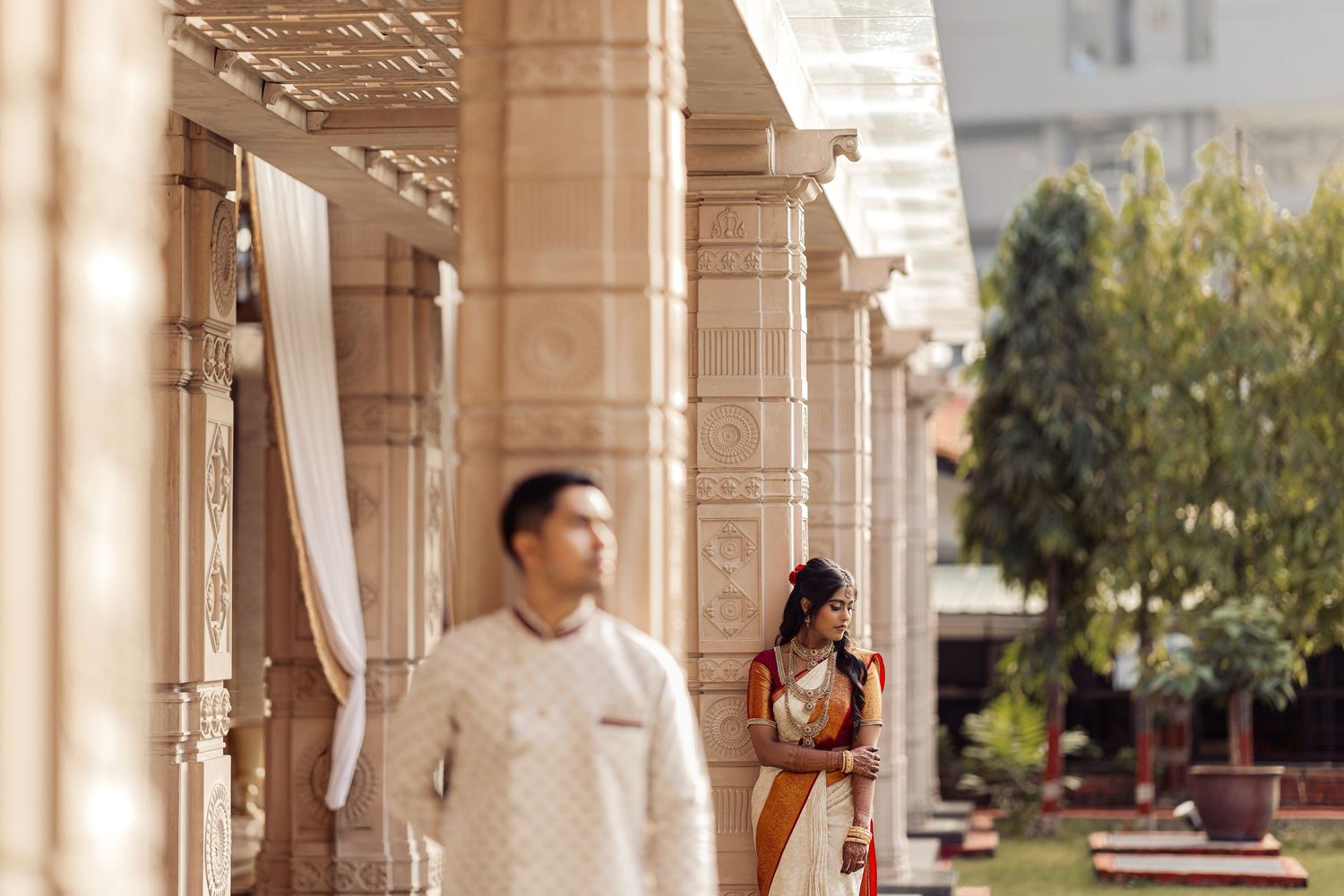 Indian couple in traditional attire posing at ornate temple corridor with elegant stone pillars.