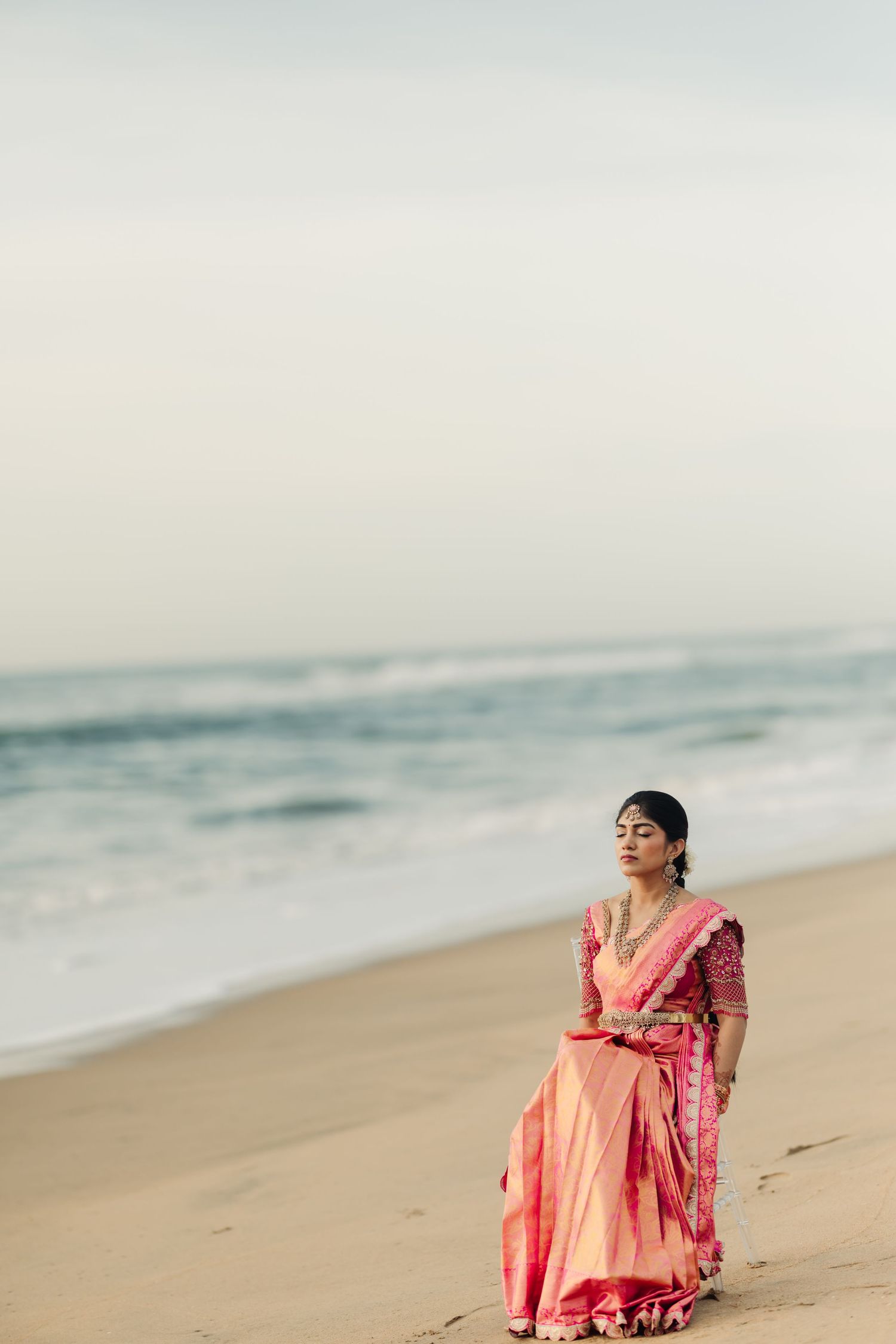 Person in pink and peach traditional attire poses gracefully on a serene beach.