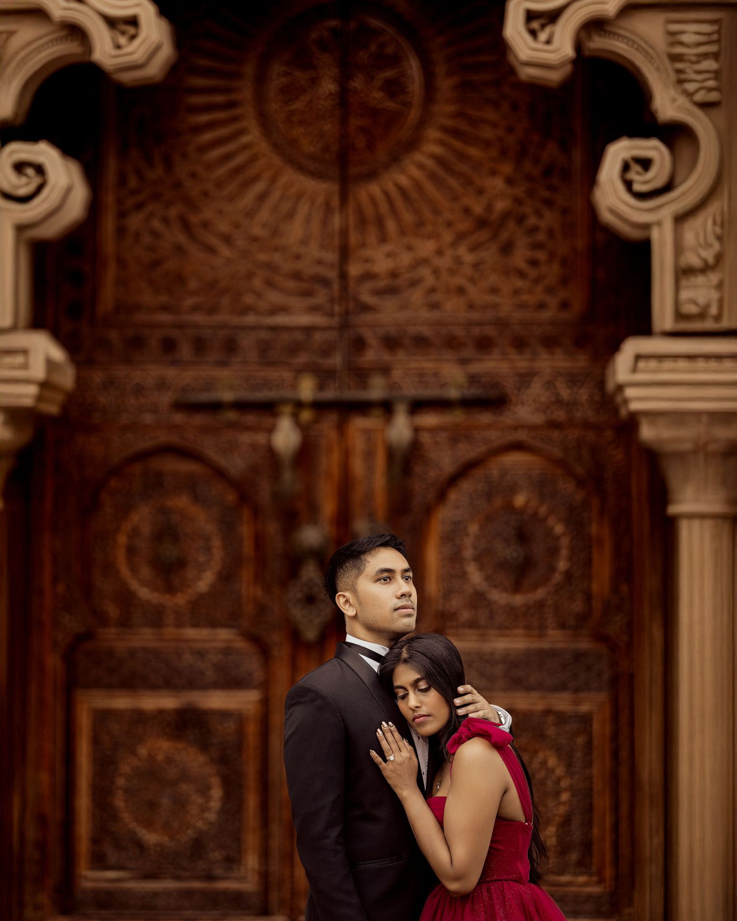 Couple in formal attire embracing in an ornate ballroom with elegant architectural details and warm lighting.