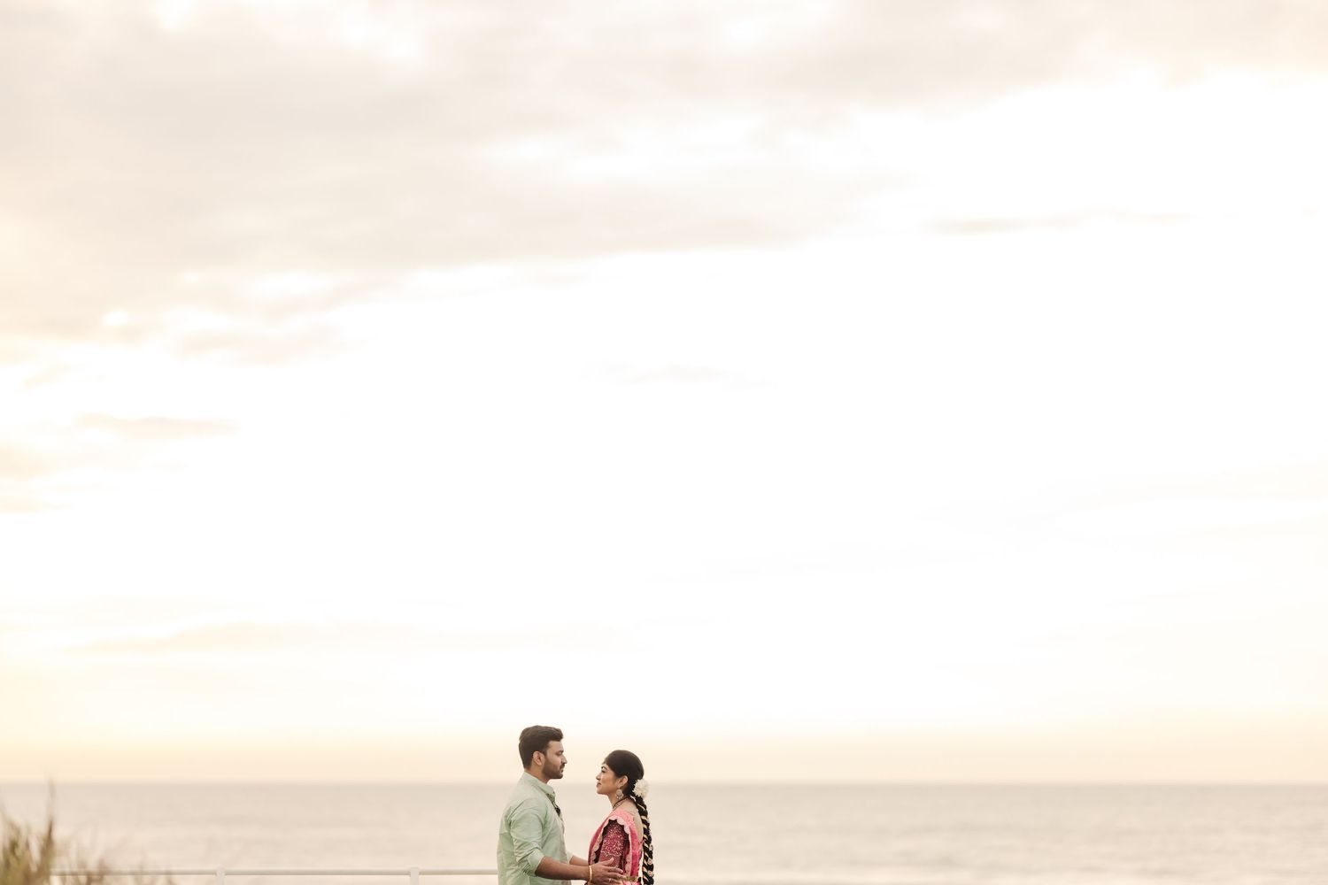 Two people stand together on a beach at sunset against a soft pink sky and ocean backdrop.