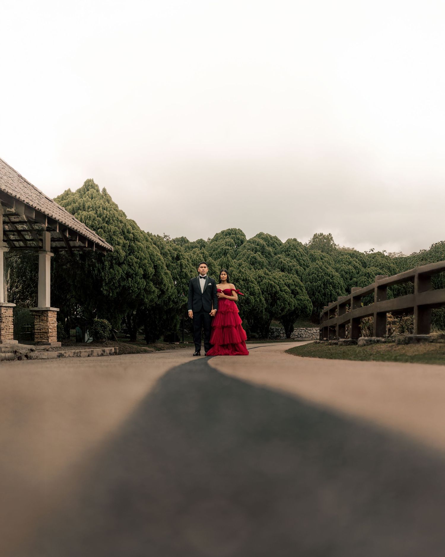 Couple in formal attire, man in black tuxedo and woman in red gown, standing on a driveway surrounded by lush trees.