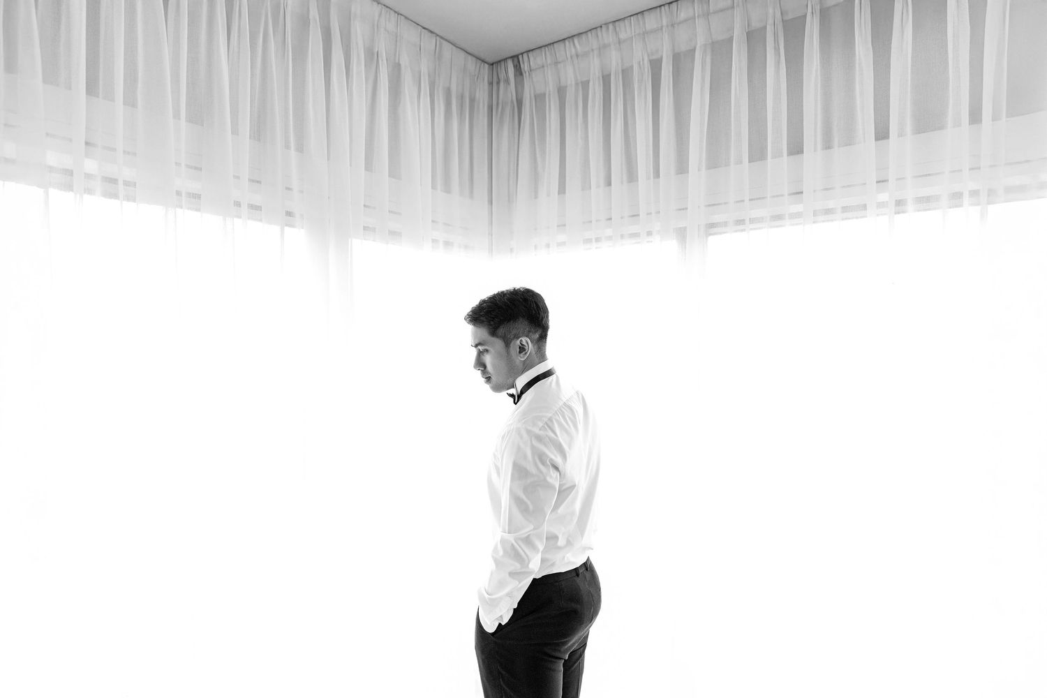 Black and white photo of a groom in a white dress shirt adjusting his bow tie before the wedding ceremony.