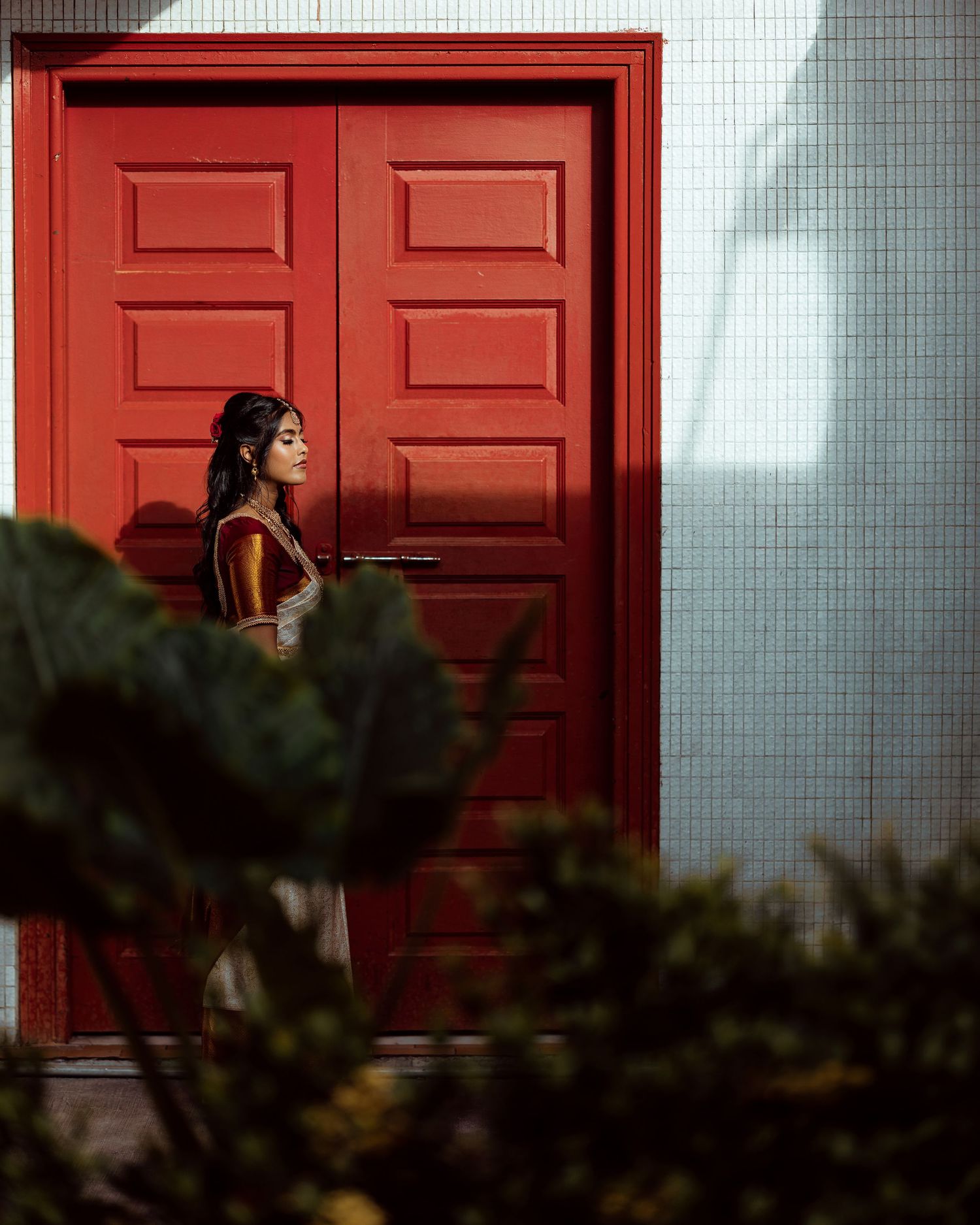 Woman in traditional attire standing before a bold red double door, framed by lush green plants in soft focus.