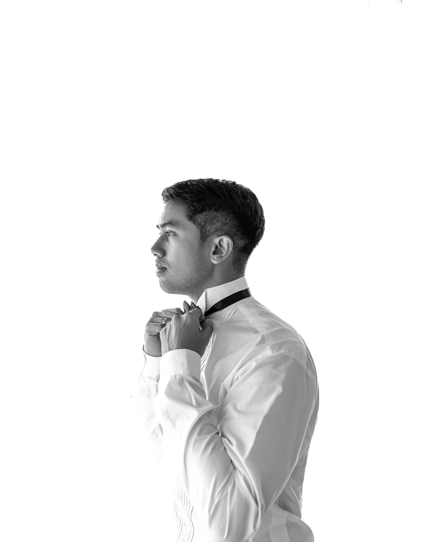 Black and white photo of a young man in a white dress shirt adjusting his bow tie, side profile view.