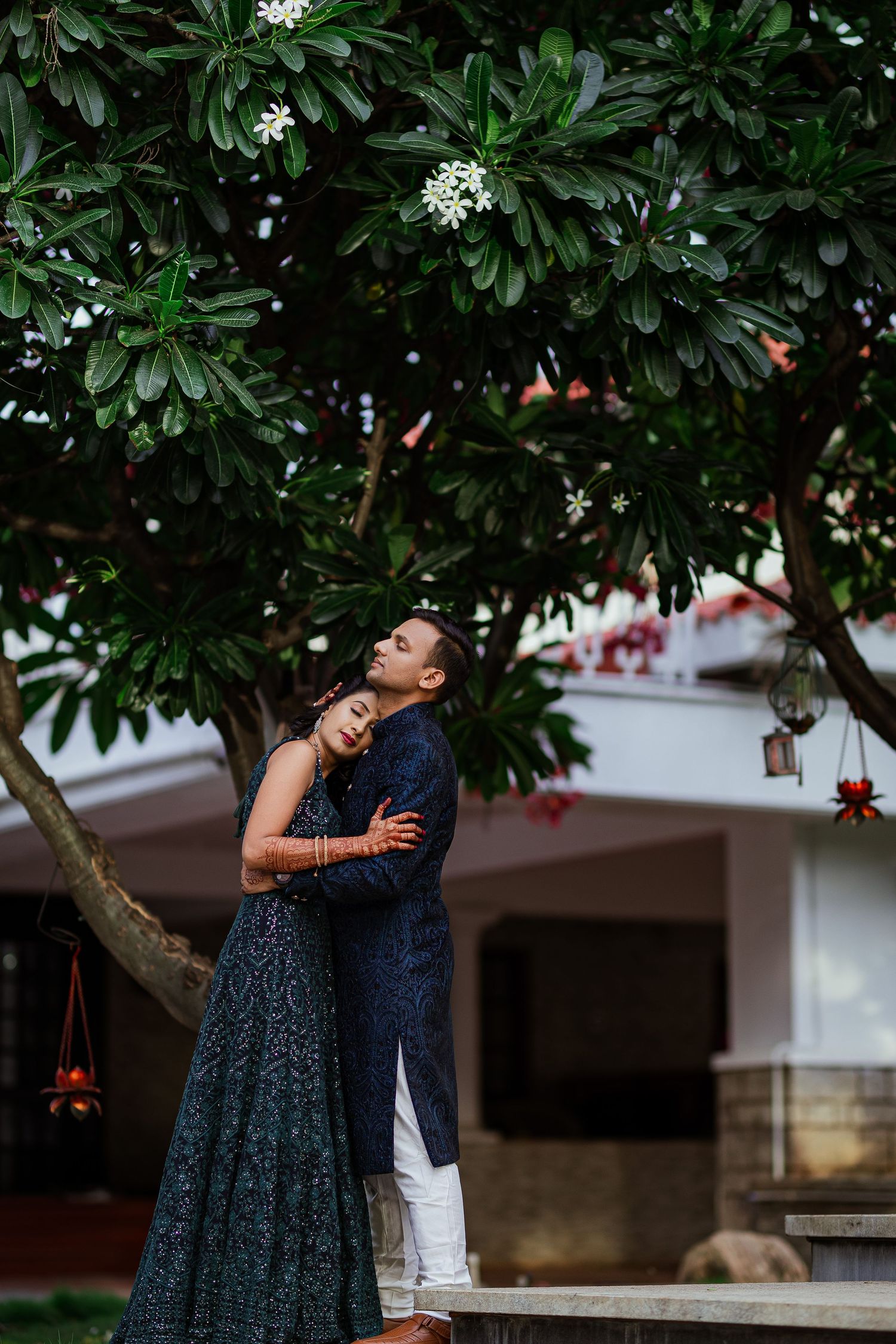 A romantic couple embraces under a flowering tree in formal attire during an outdoor photoshoot.