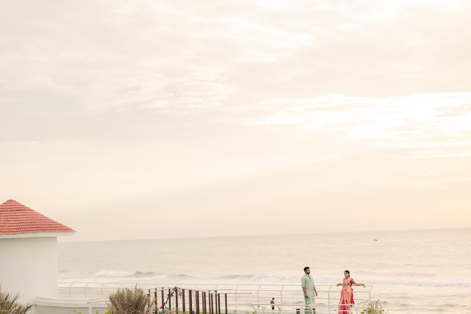 Two figures stand on a beach at sunset near a wooden fence and red umbrella by the ocean.