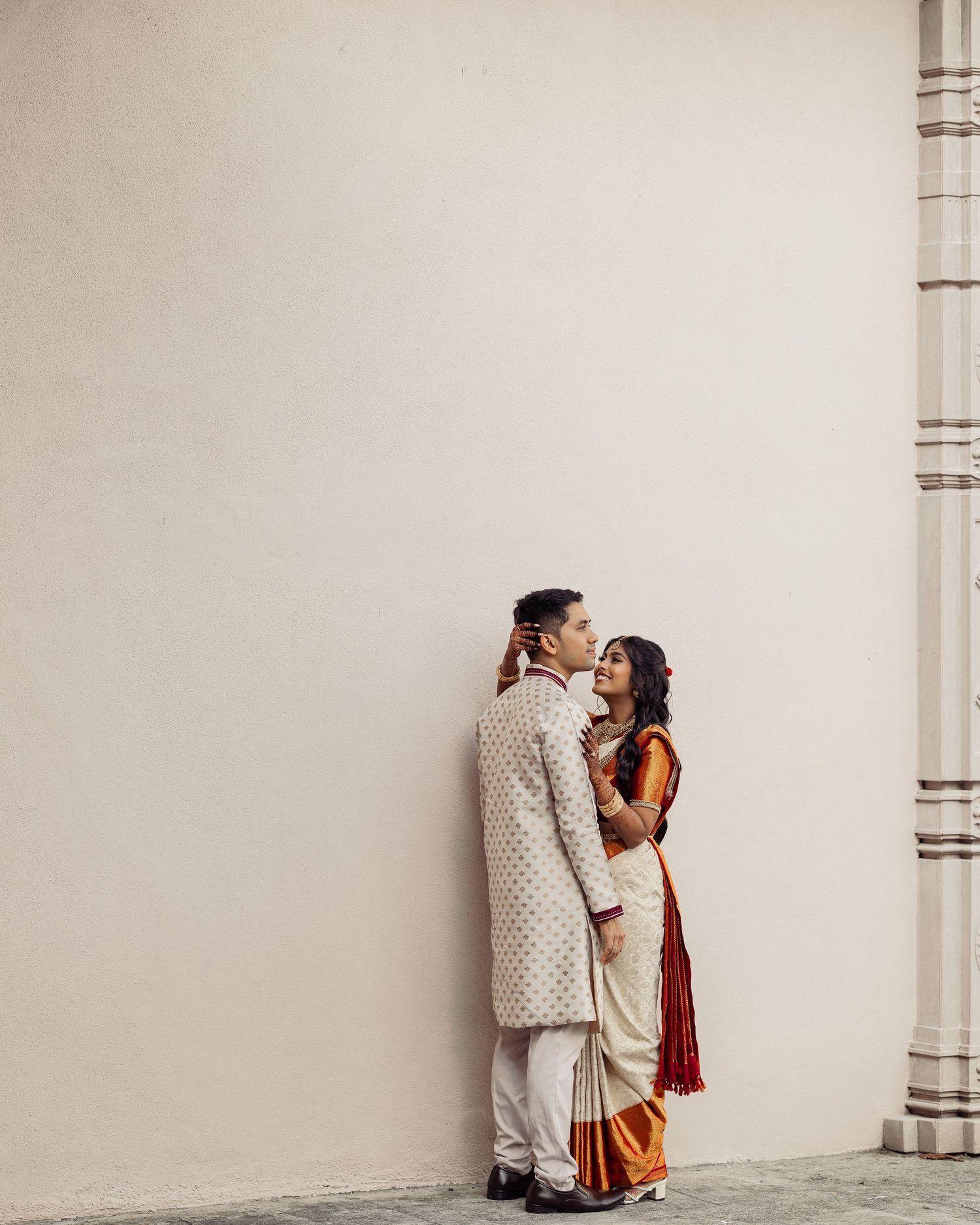 Couple in traditional Indian wedding attire embracing against a white wall, groom in sherwani and bride in colorful saree.