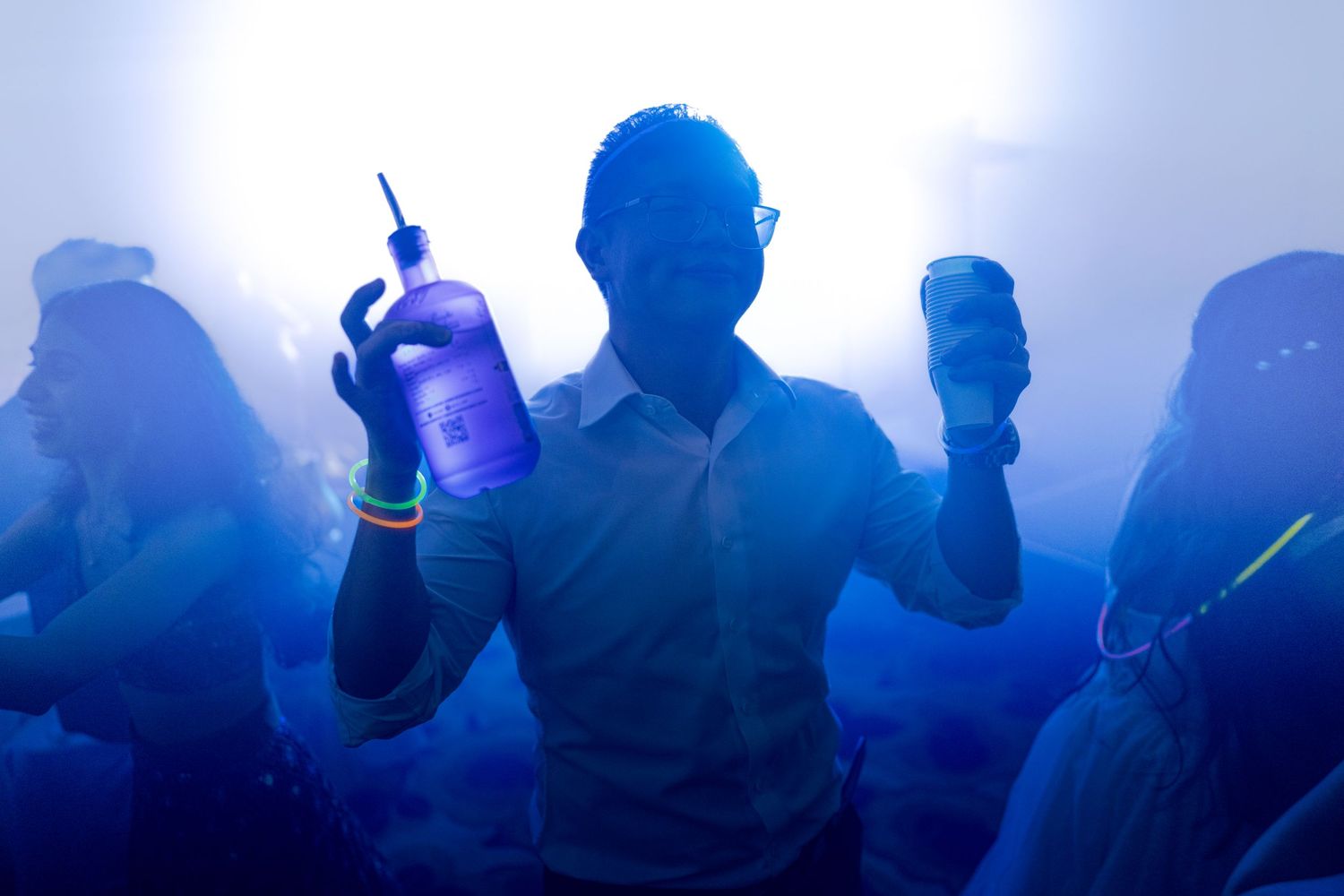 Man holding glowing bottle and can at a blue-lit nightclub party with fog and dancing crowd.