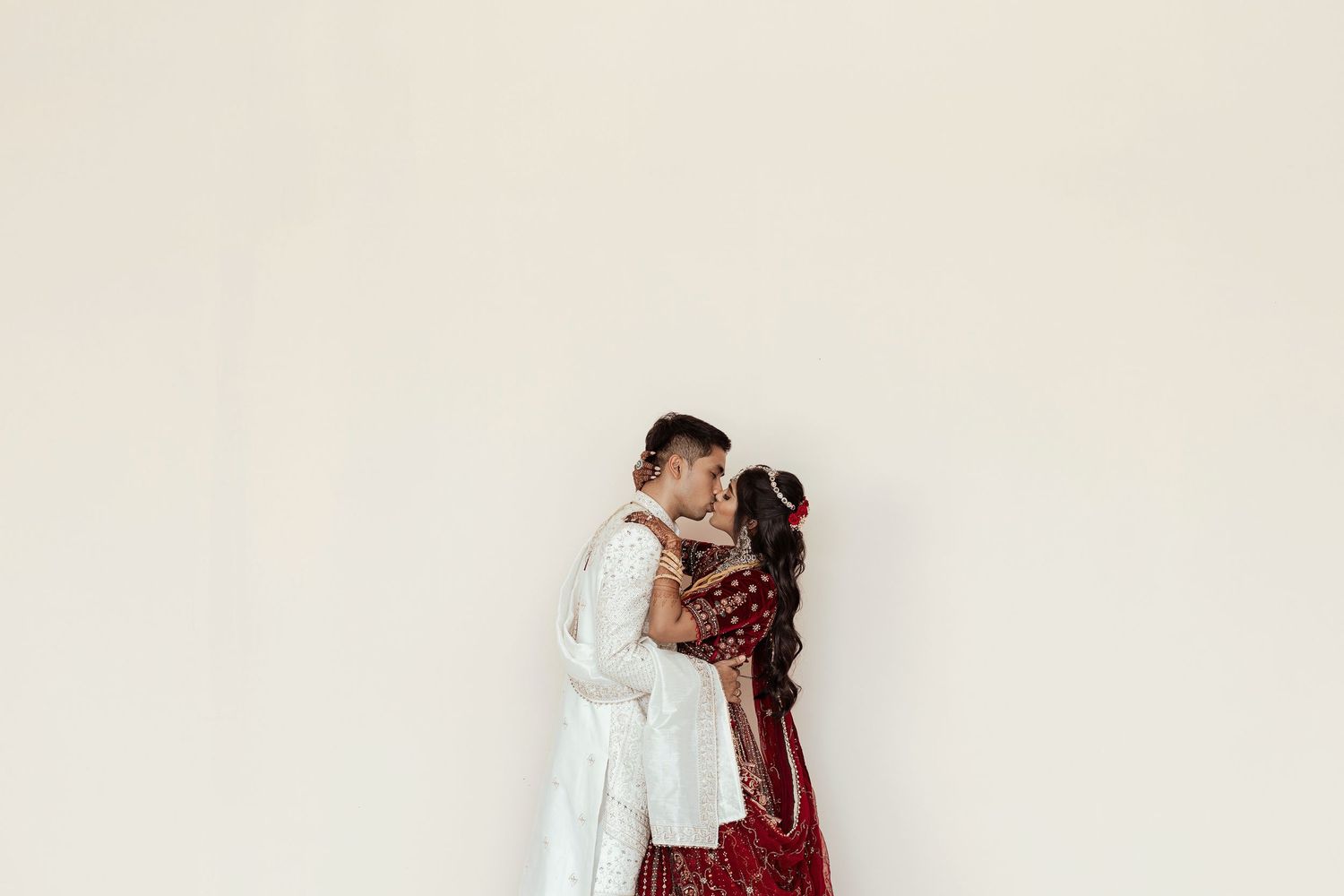 A couple in traditional South Asian wedding attire shares a romantic kiss, wearing white sherwani and red bridal lehenga.