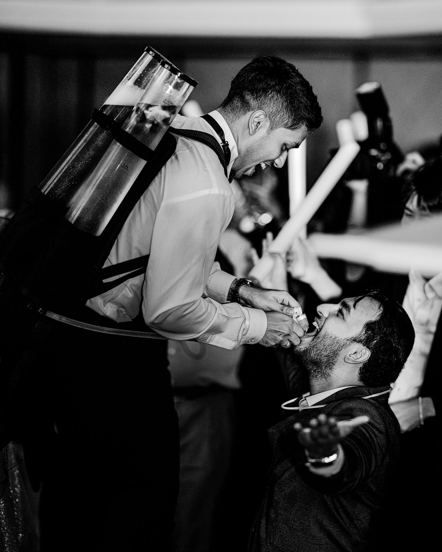 Black and white photo of a bartender pouring a drink directly into a mans mouth at a lively party event.