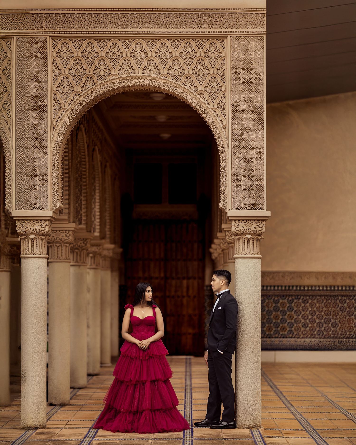 A couple poses in an ornate Moroccan-style palace corridor with arched doorways and intricate tile patterns.