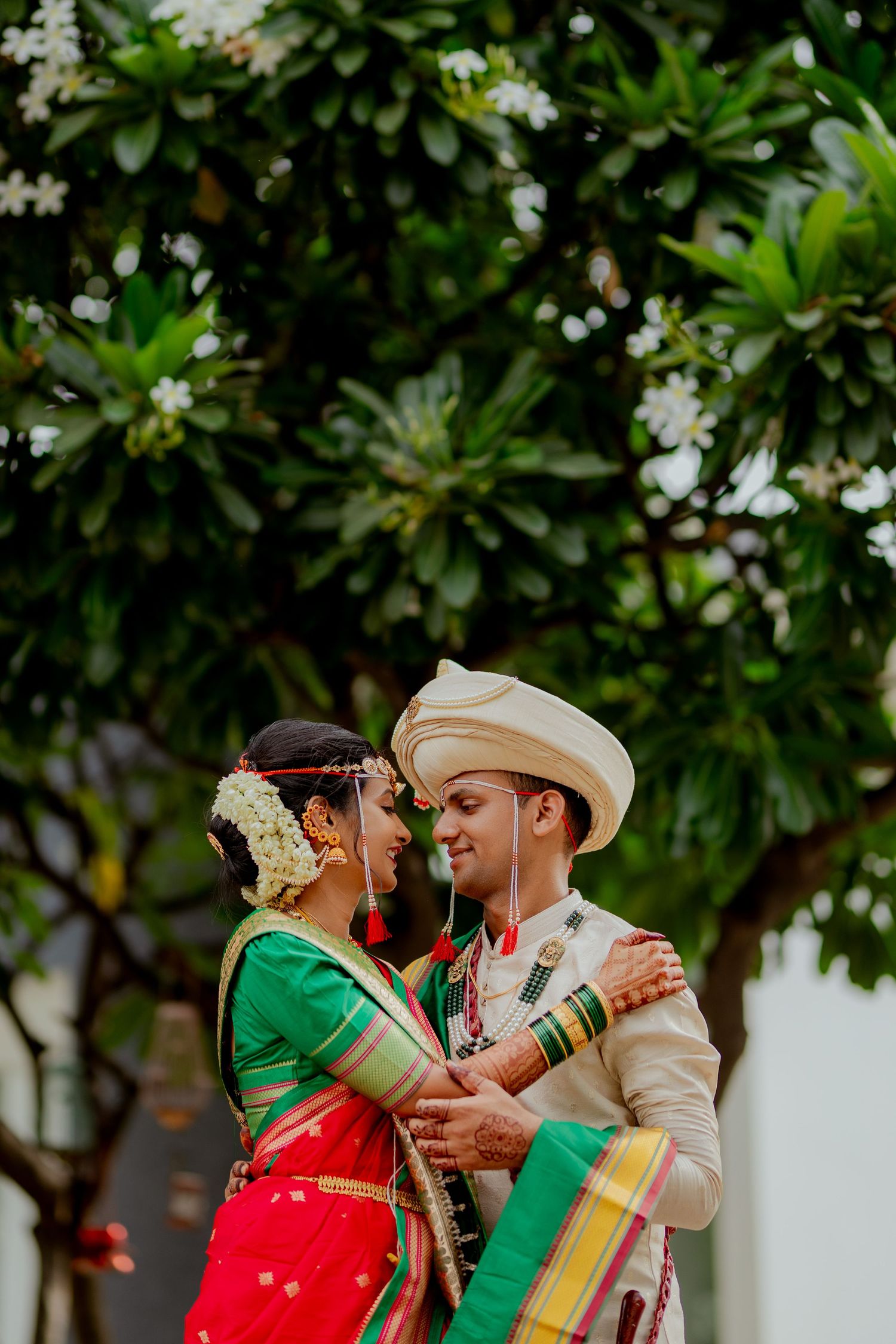 A couple in traditional Indian wedding attire embrace during their outdoor ceremony under flowering trees.