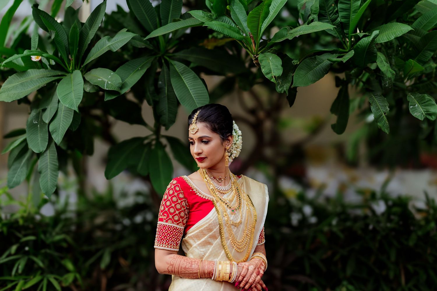 A bride in a traditional South Indian wedding saree poses gracefully against lush green foliage in an outdoor setting.