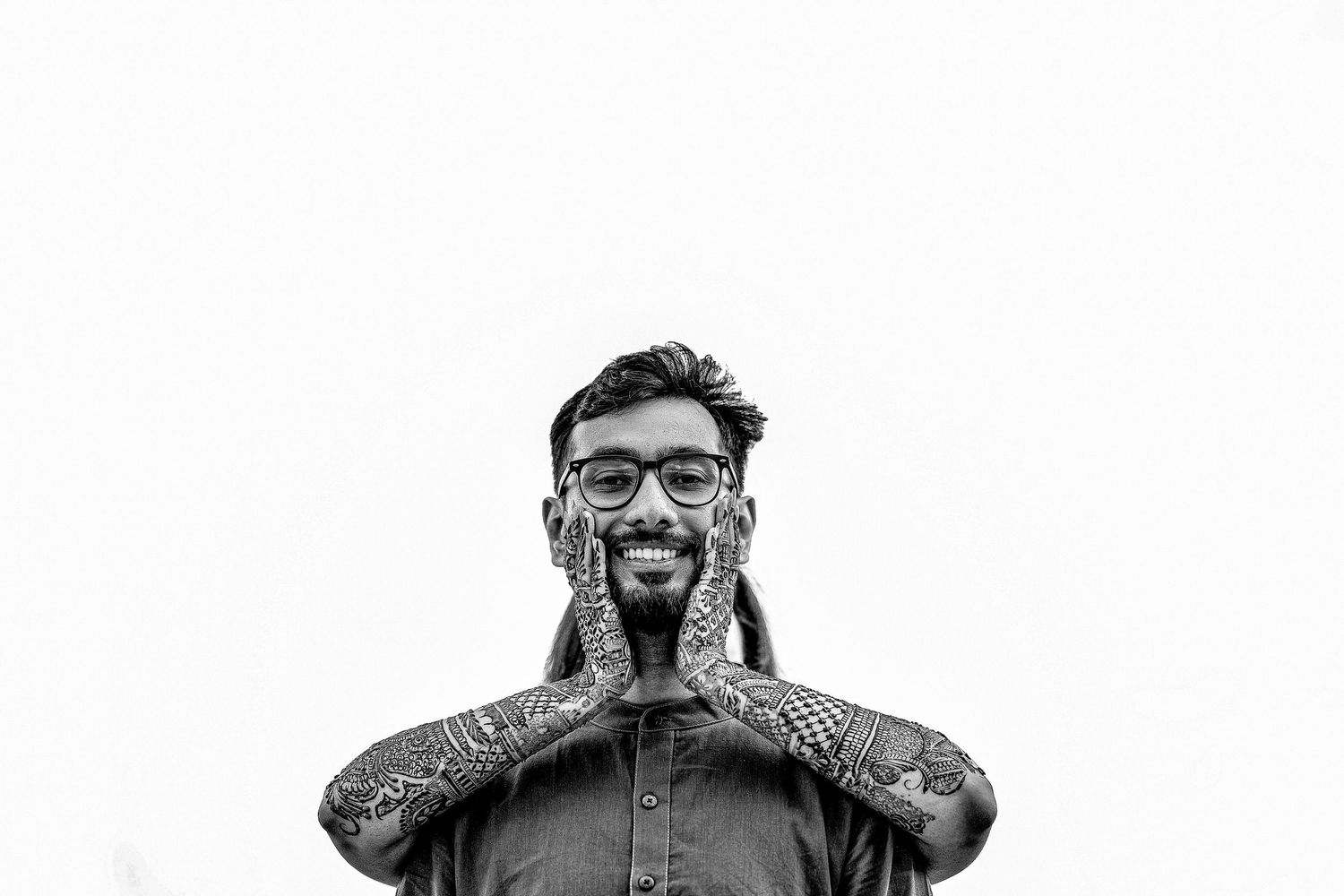 A person wearing glasses and a denim shirt smiles while holding their hands to their face in a black and white photo.