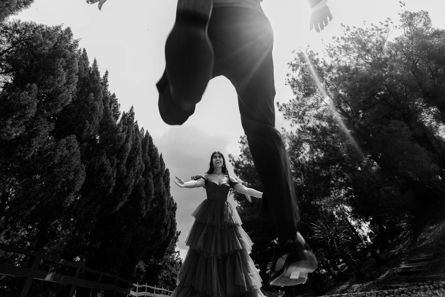Black and white photo of bride in layered gown with groom jumping above her in a dramatic low-angle outdoor shot.