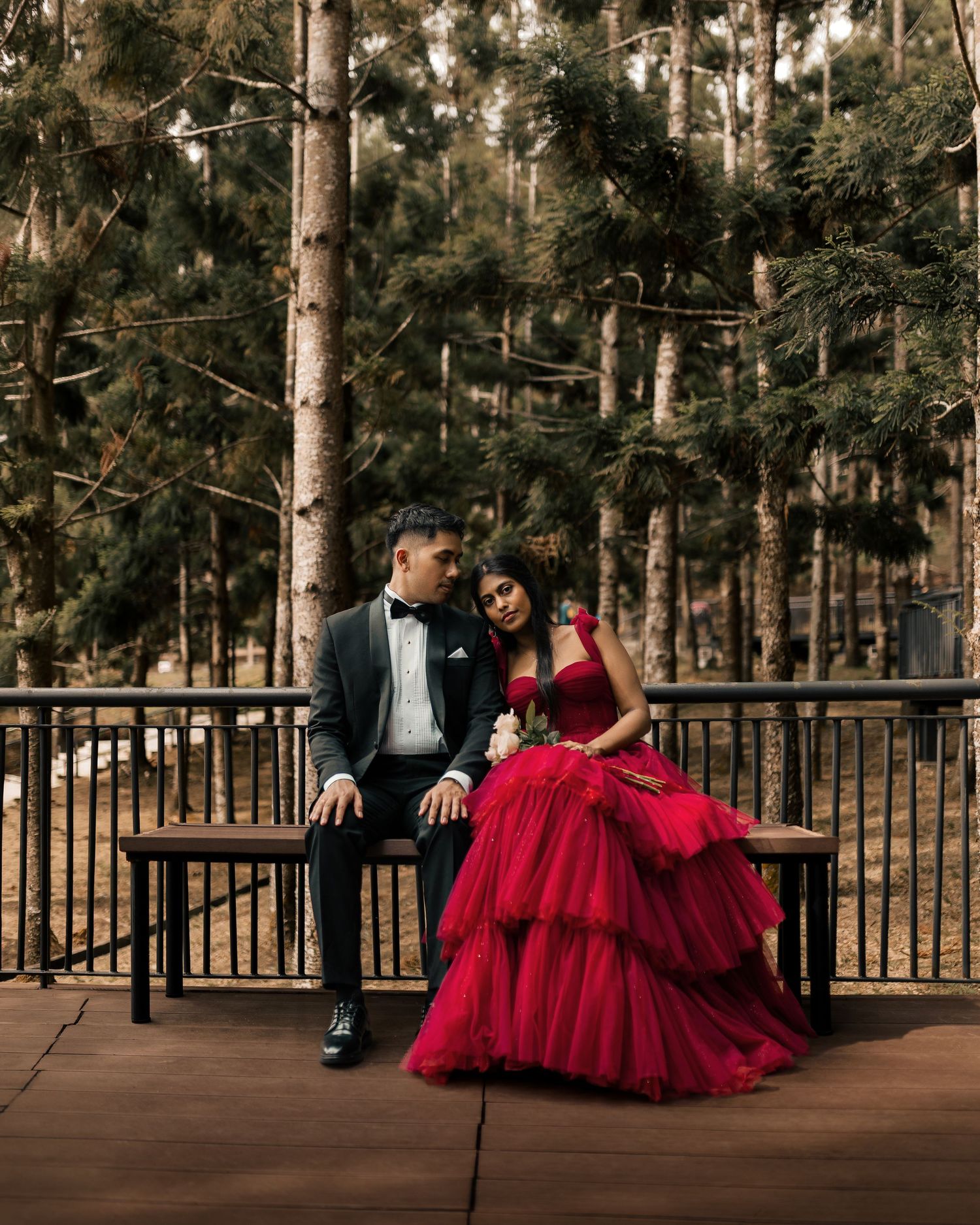 Couple in formal wear sitting on bench in forest, man in tuxedo and woman in red ball gown.