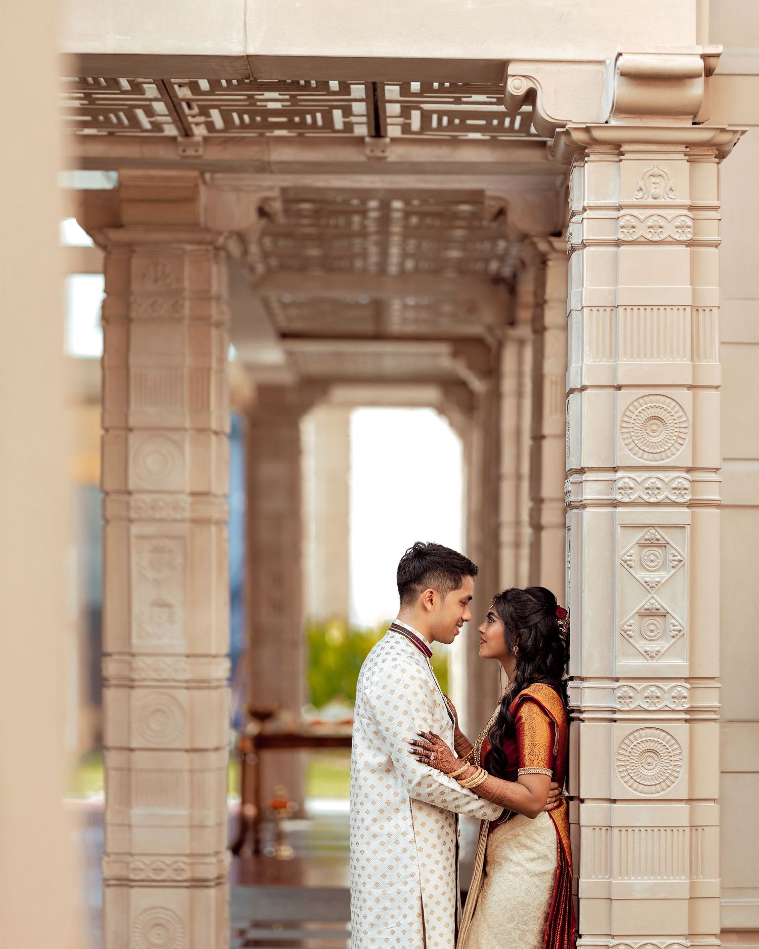 Indian couple in traditional attire sharing a romantic moment between ornate white temple columns.