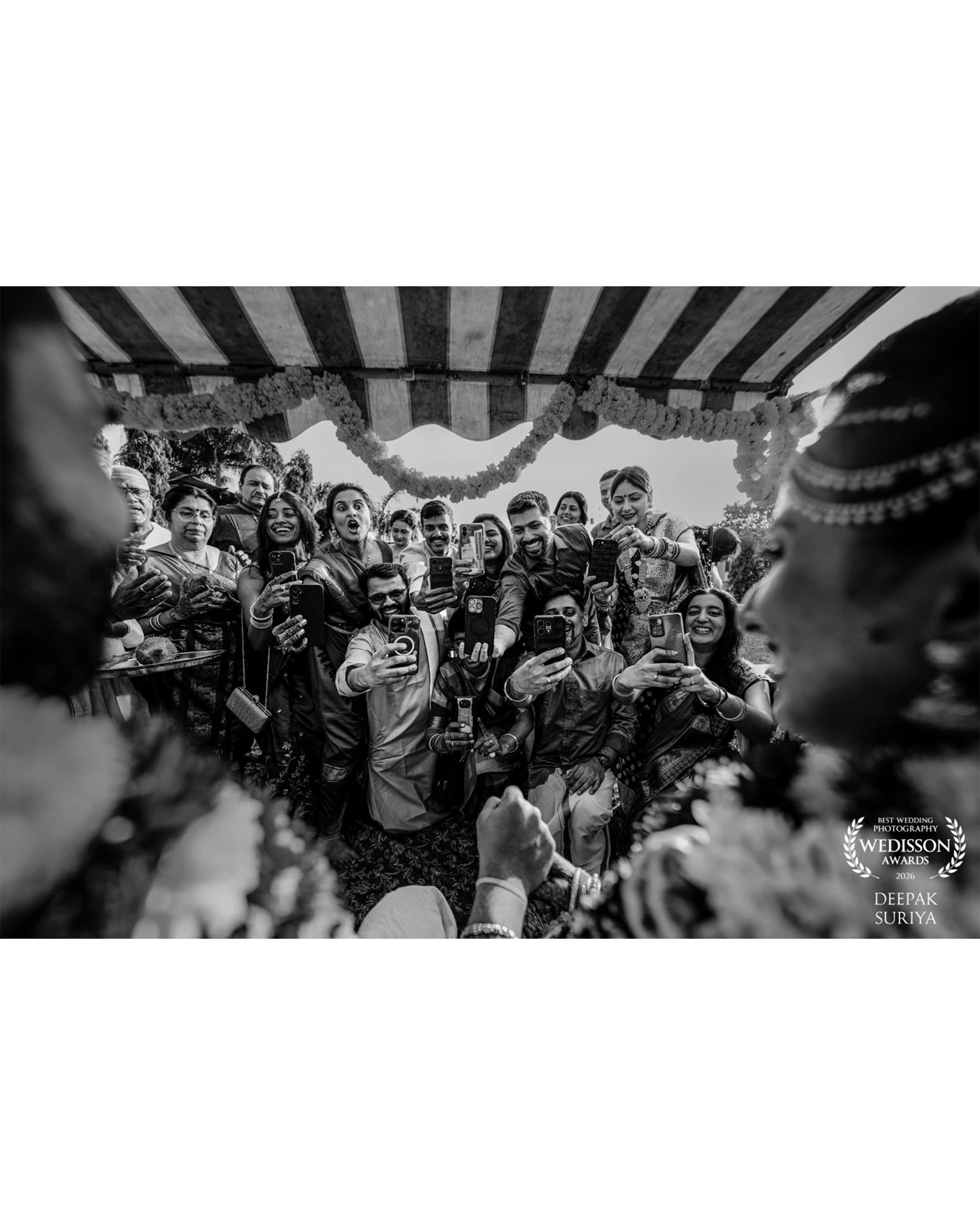 Black and white wedding photo of crowd capturing moments on phones, framed by floral decorations and striped canopy.