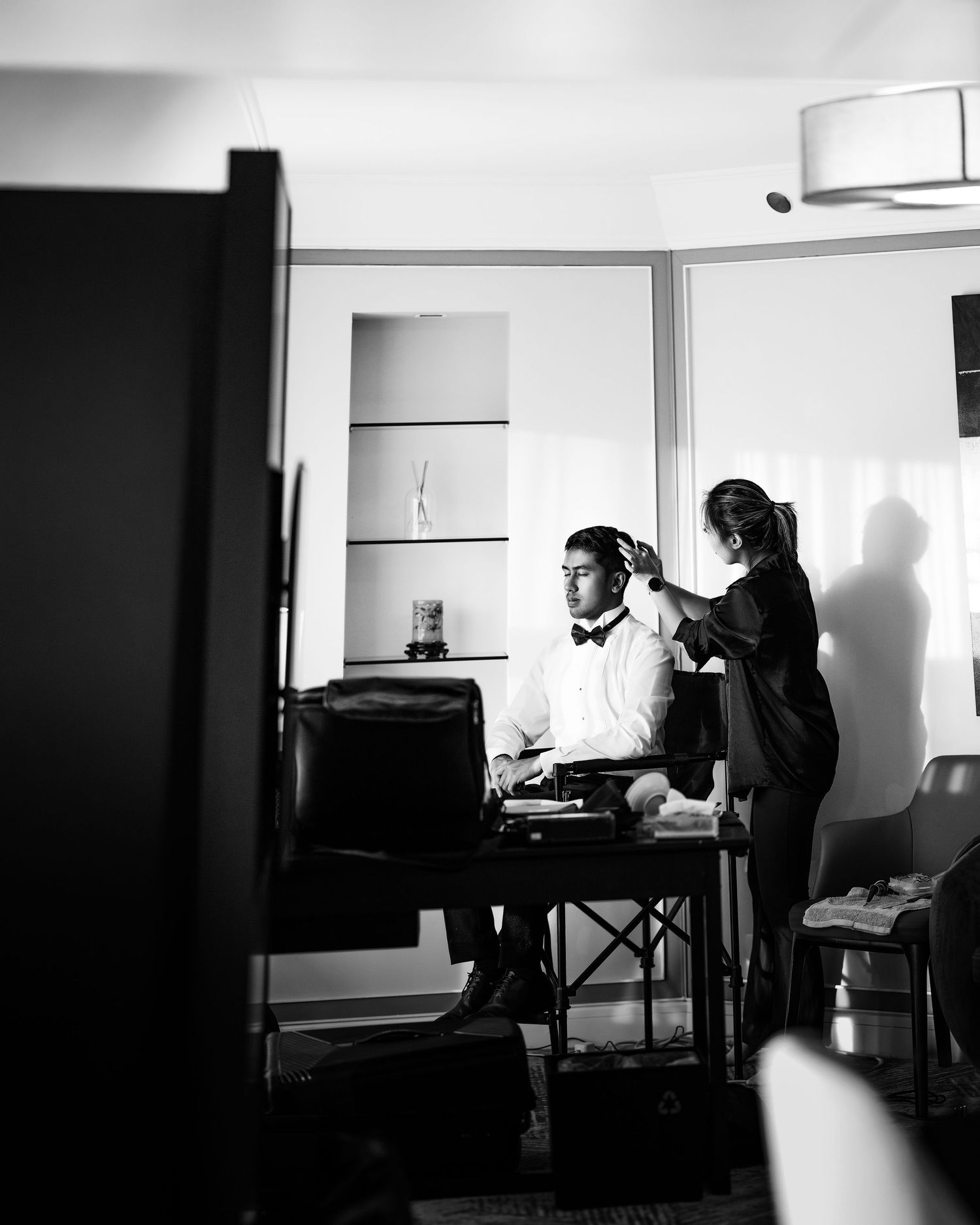 Black and white photo of a groom in bow tie getting ready, assisted by a stylist in an elegant room.