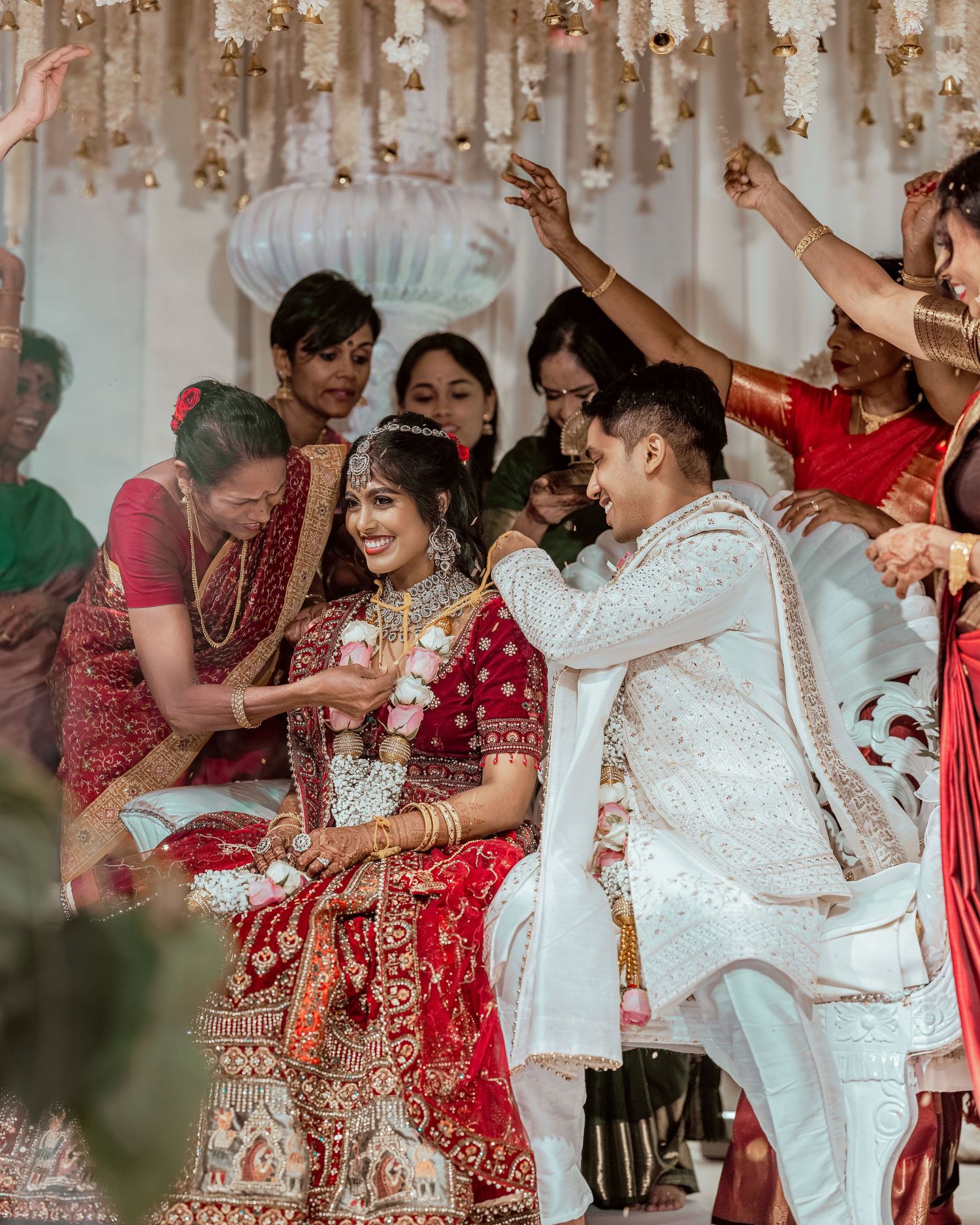 Indian bride in red lehenga and groom in white sherwani during traditional Hindu wedding ceremony with guests celebrating.