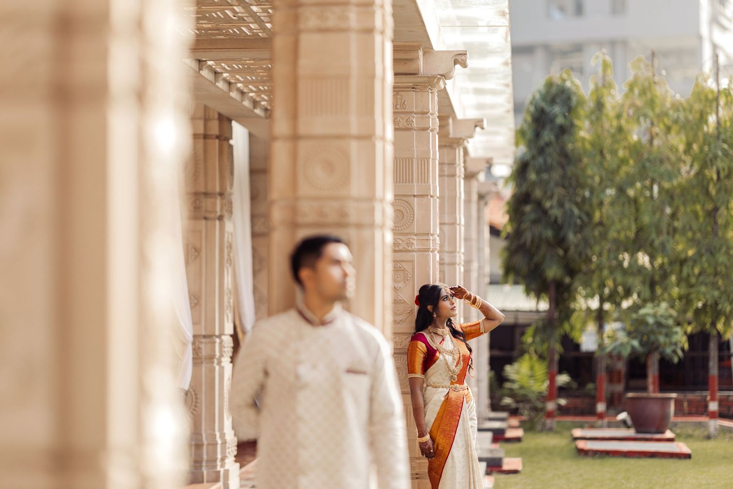 Couple in traditional Indian attire posing at ornate temple columns, man in white kurta, woman in orange saree.