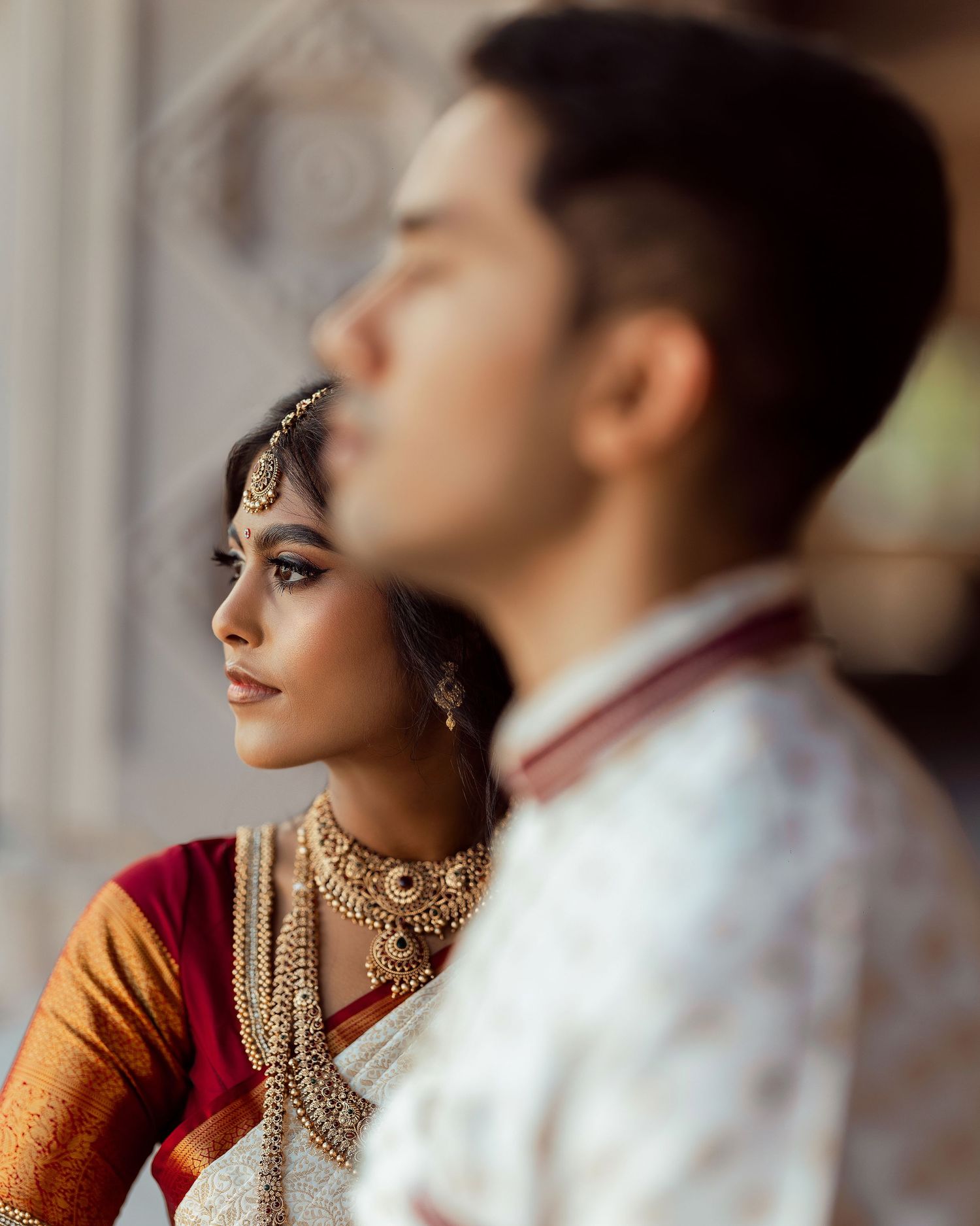Indian bride in red and gold saree with layered gold jewelry stands beside groom in white sherwani during wedding ceremony.