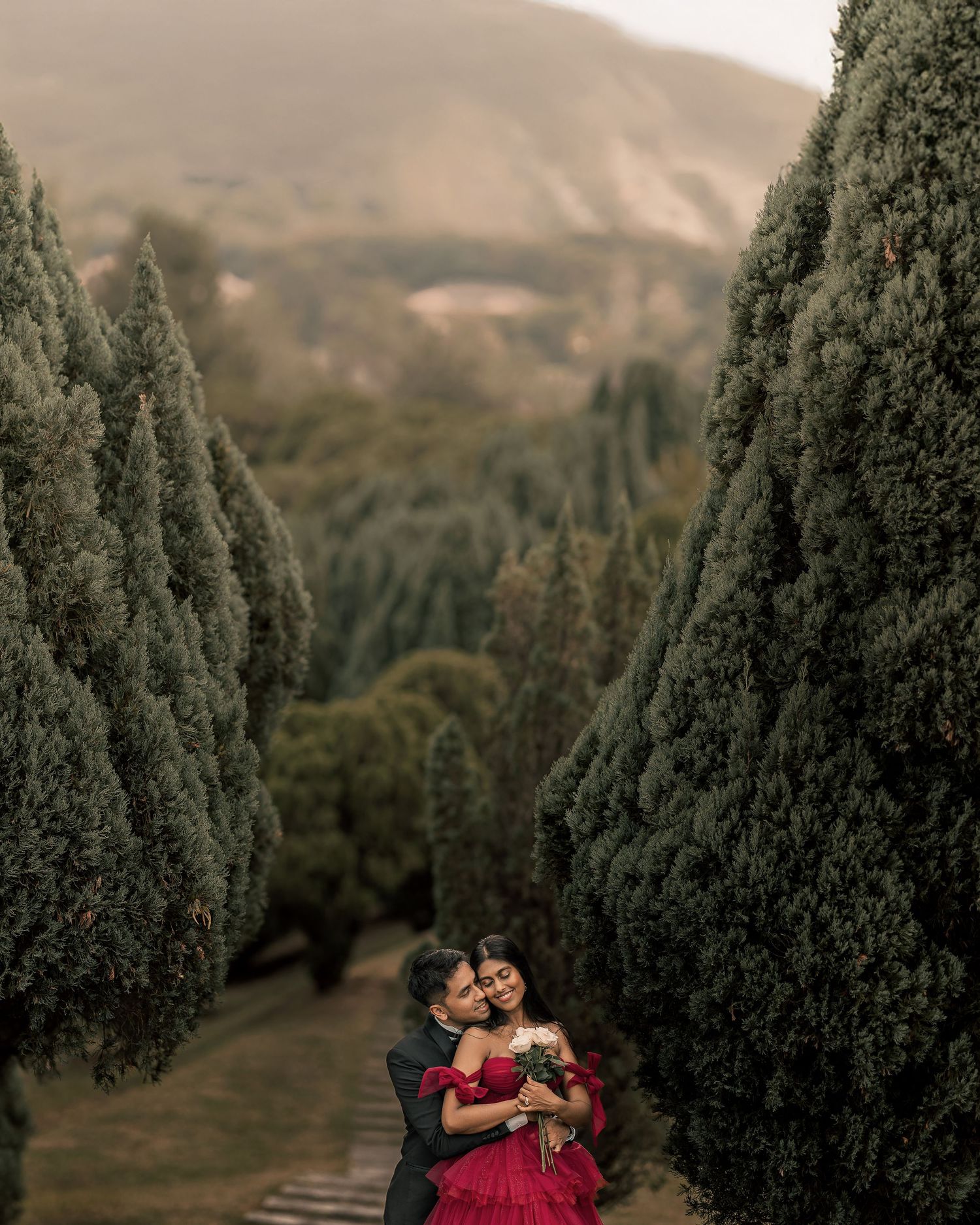 Couple embracing in a romantic alley of tall cypress trees, woman in red gown holding bouquet, mountains in background.