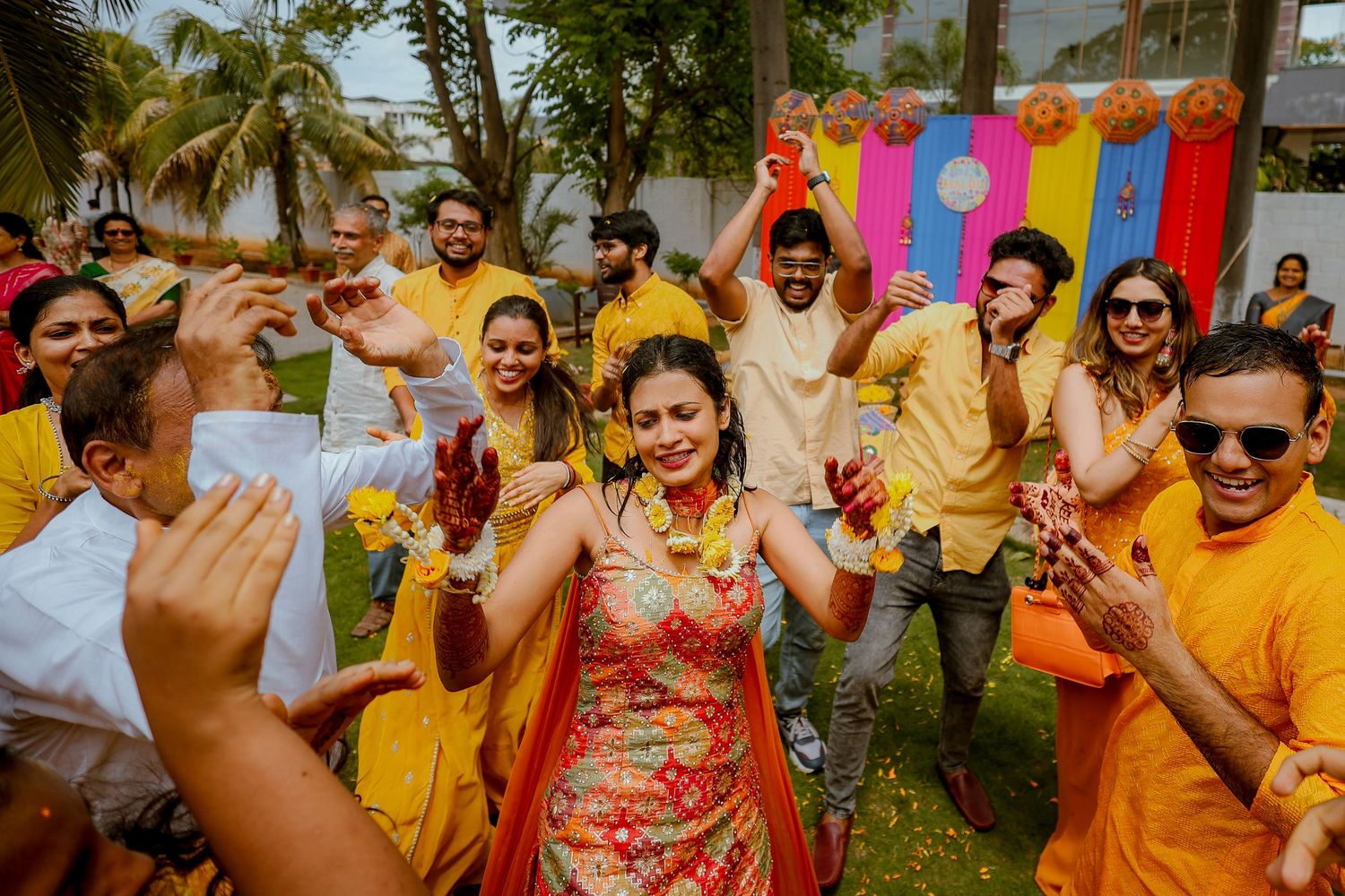 Colorful celebration at an outdoor Indian wedding ceremony with guests wearing vibrant yellow and red attire dancing joyfully.
