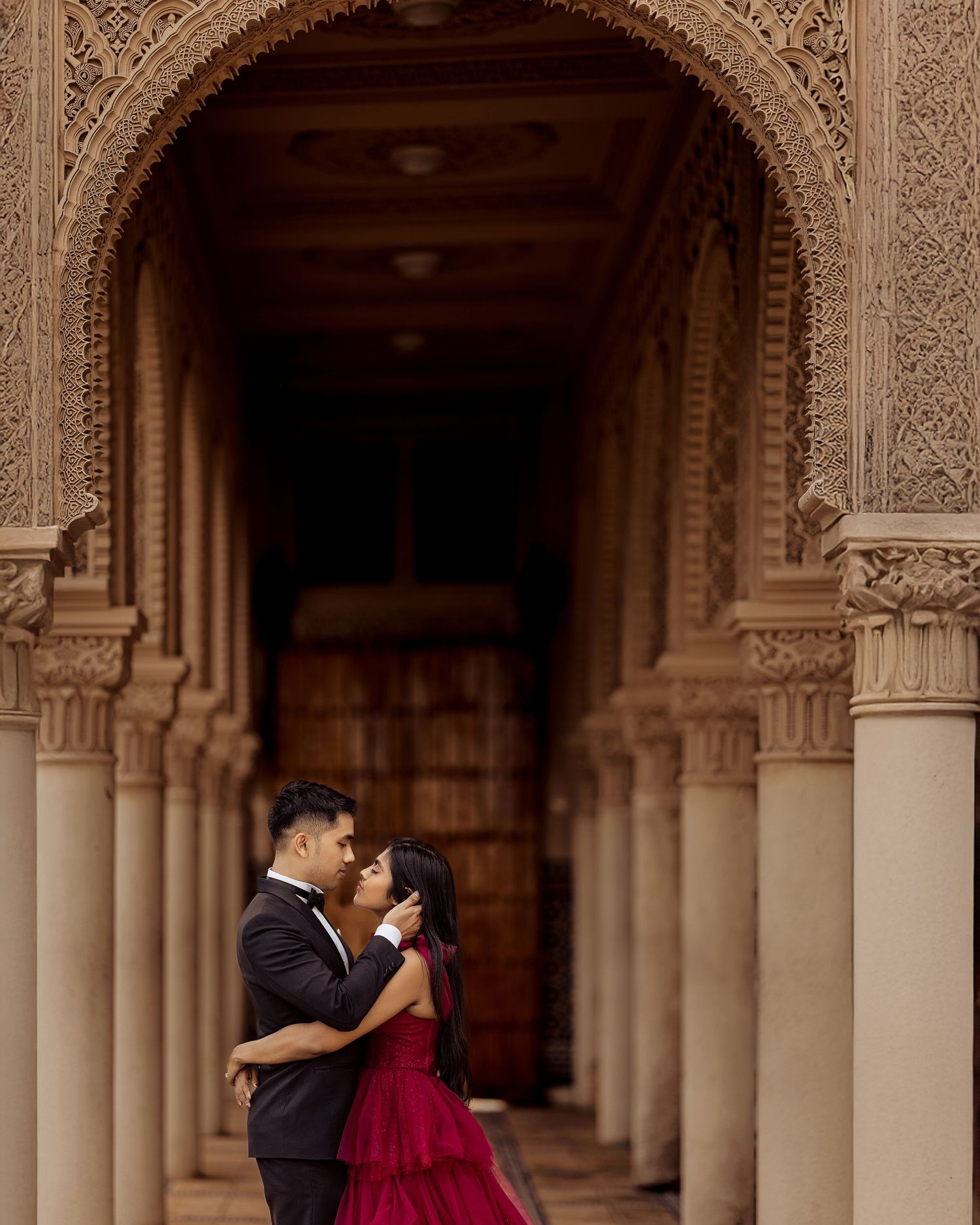 Couple embraces in formal attire against ornate archways and columns of historic architecture.
