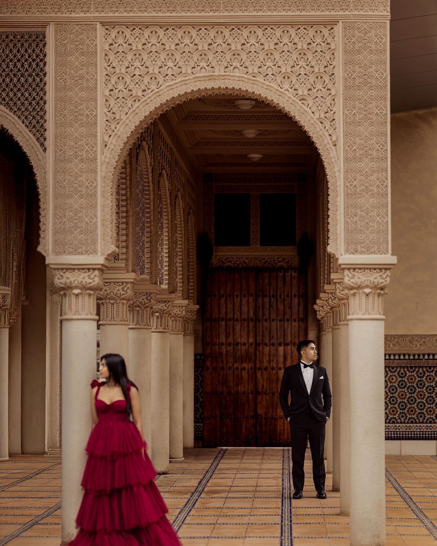 Elegant couple poses in formal attire against ornate Moorish architecture with arches and intricate patterns.