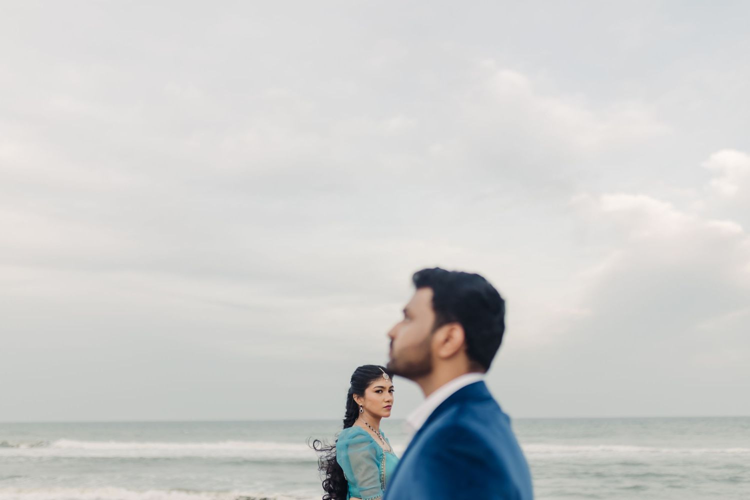 A couple stands together on a beach looking out at the ocean during an overcast day.