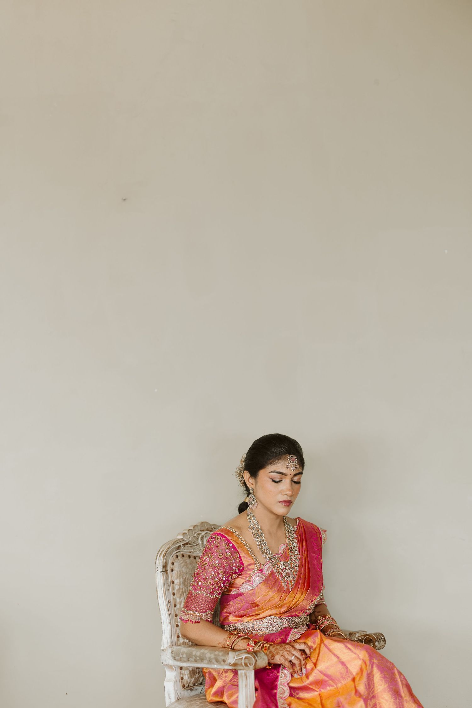 A figure in a vibrant orange and pink sari sits thoughtfully in a white chair against a neutral wall.