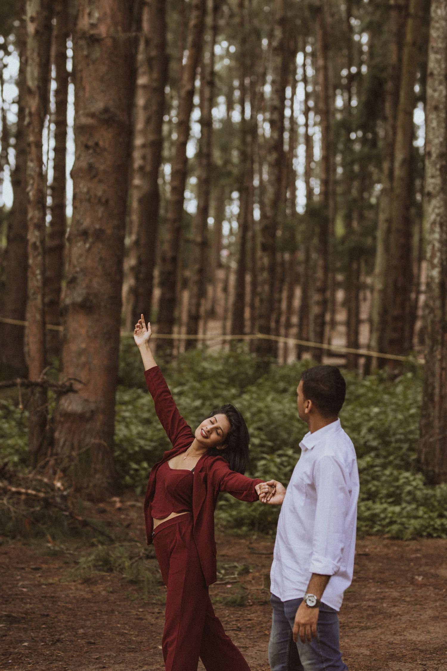 A couple poses playfully in a pine forest, with one person wearing burgundy and the other in white.