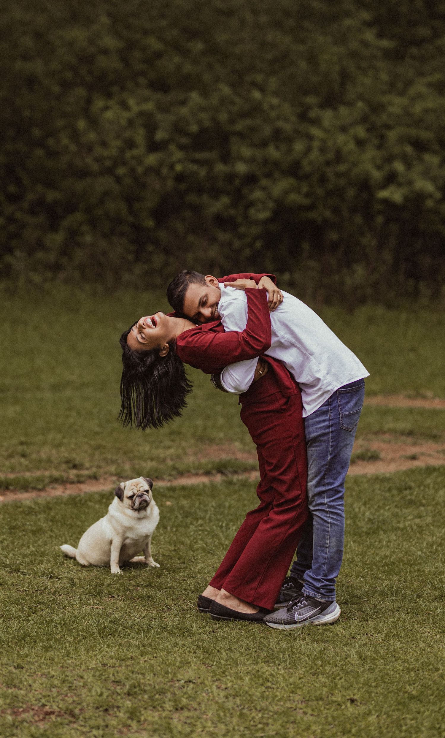 A person in a red jacket bends backwards playfully on grass while a pug dog sits nearby watching.