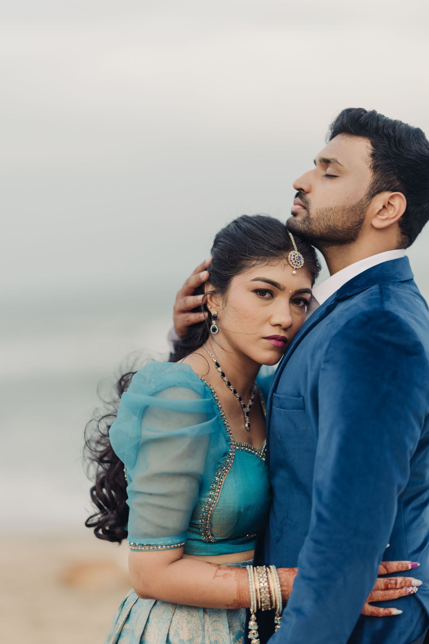 A romantic couple in traditional South Asian attire embrace on a misty beach, with her in turquoise and him in navy blue.