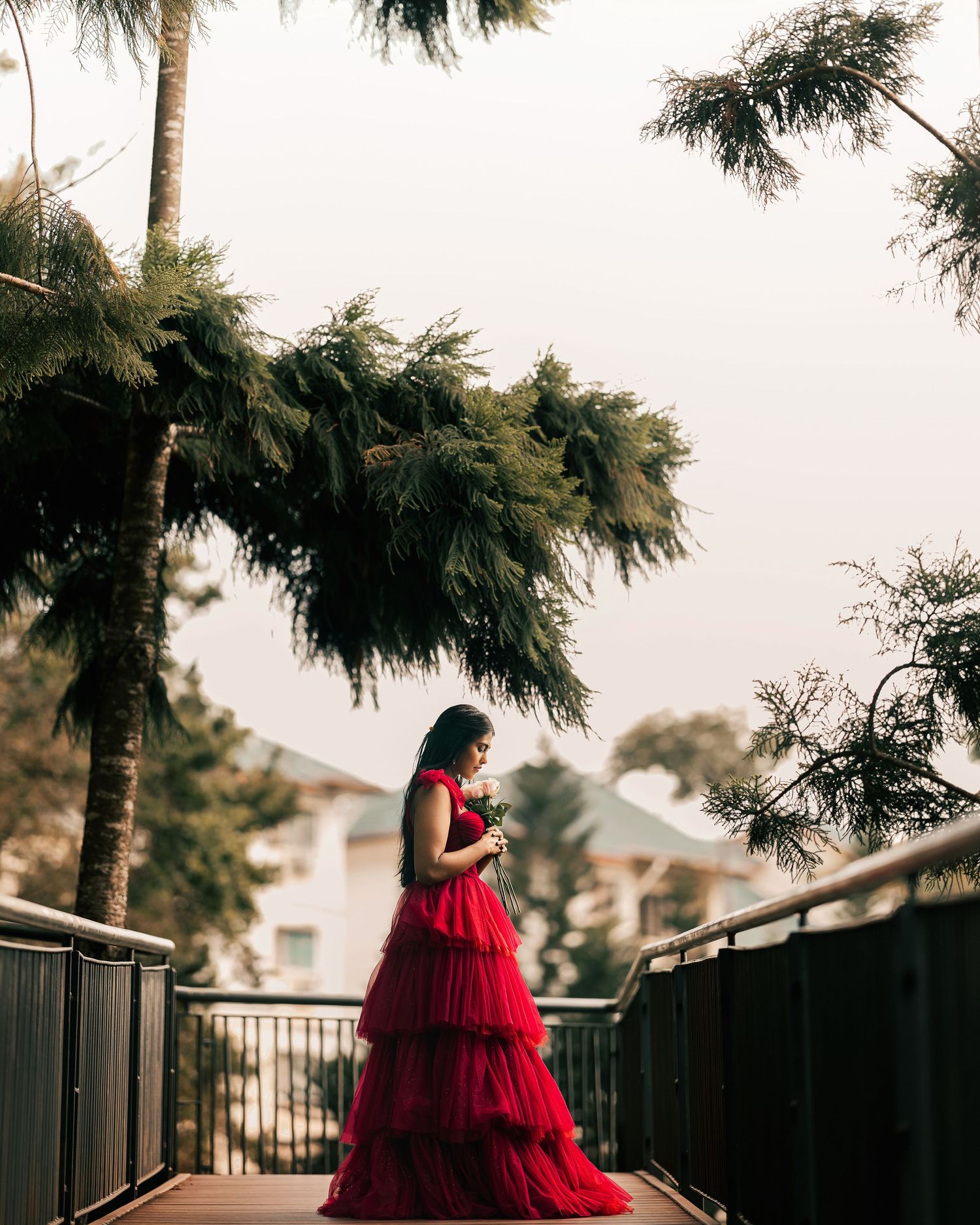 Woman in red tiered gown standing on a wooden bridge surrounded by lush green trees.