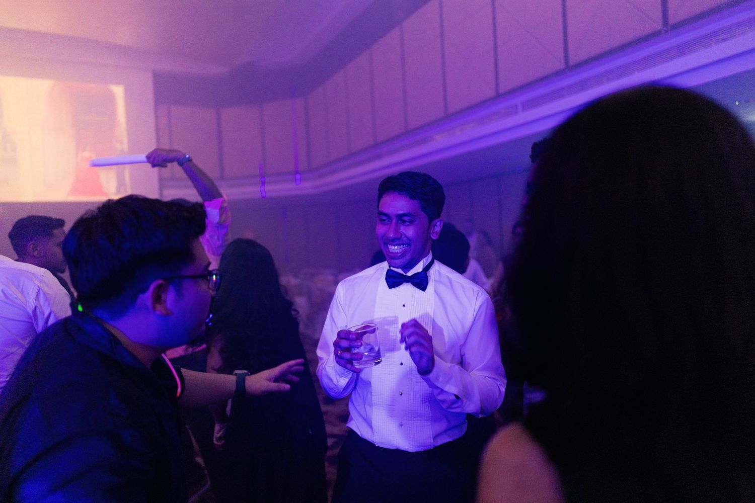 People socializing at a formal event with purple lighting, men in tuxedos mingling on a dance floor.