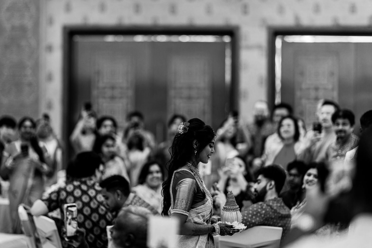 Black and white photo of a bride in a saree surrounded by guests at an Indian wedding celebration.