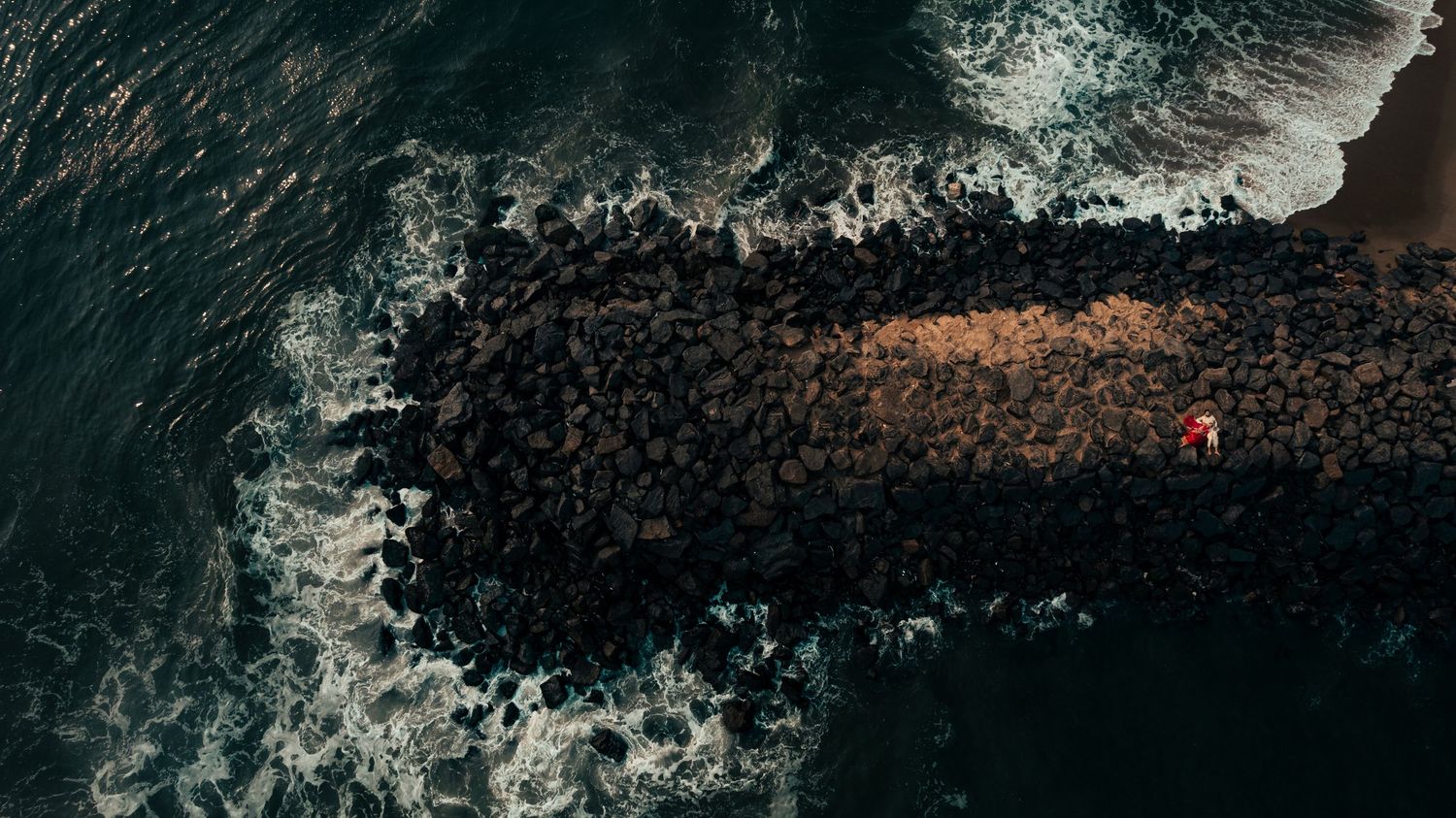Aerial view of a rocky jetty extending into dark ocean waves with a lone figure in red standing at the tip.