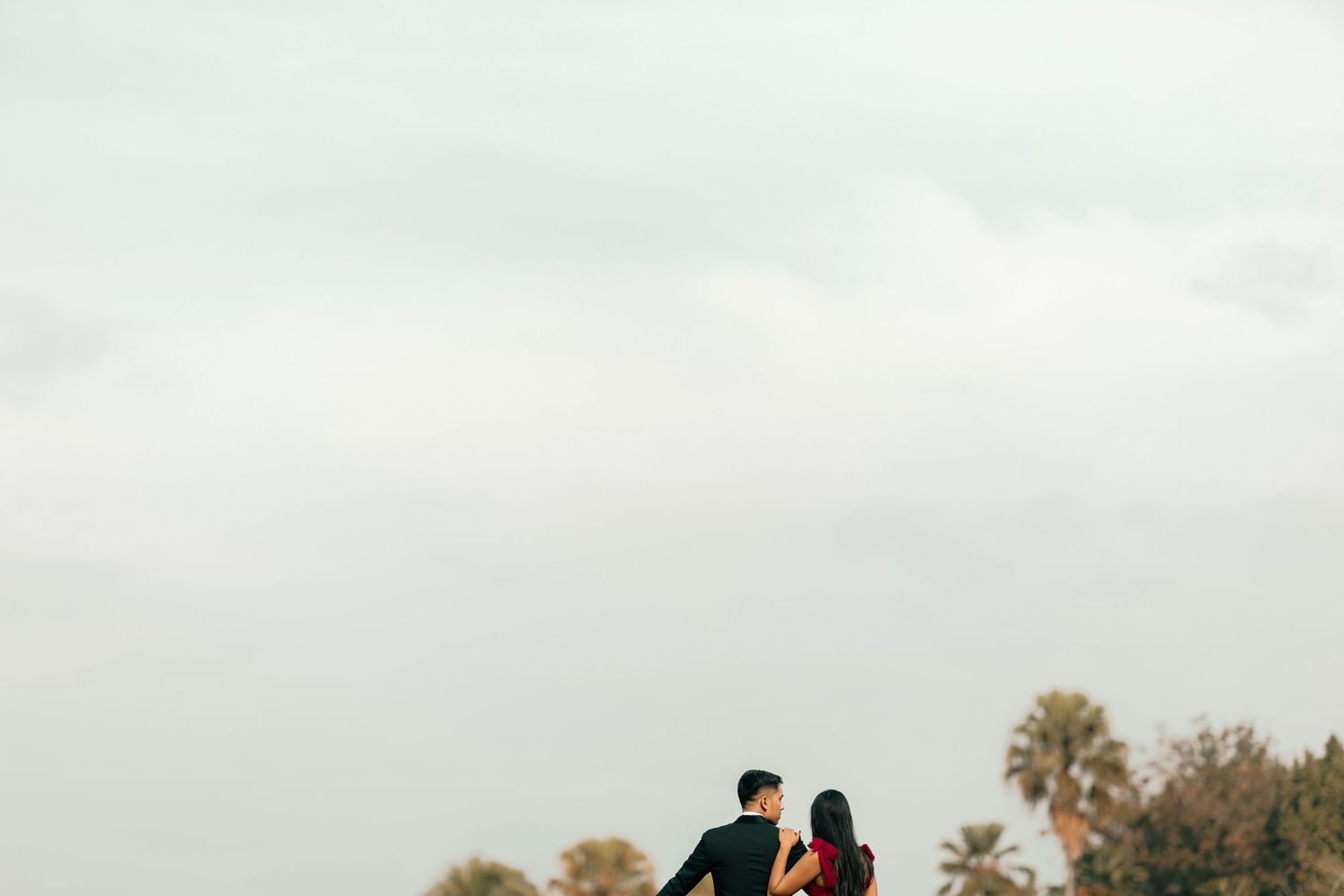 A couple stands together against a cloudy sky with palm trees, sharing a romantic moment outdoors.