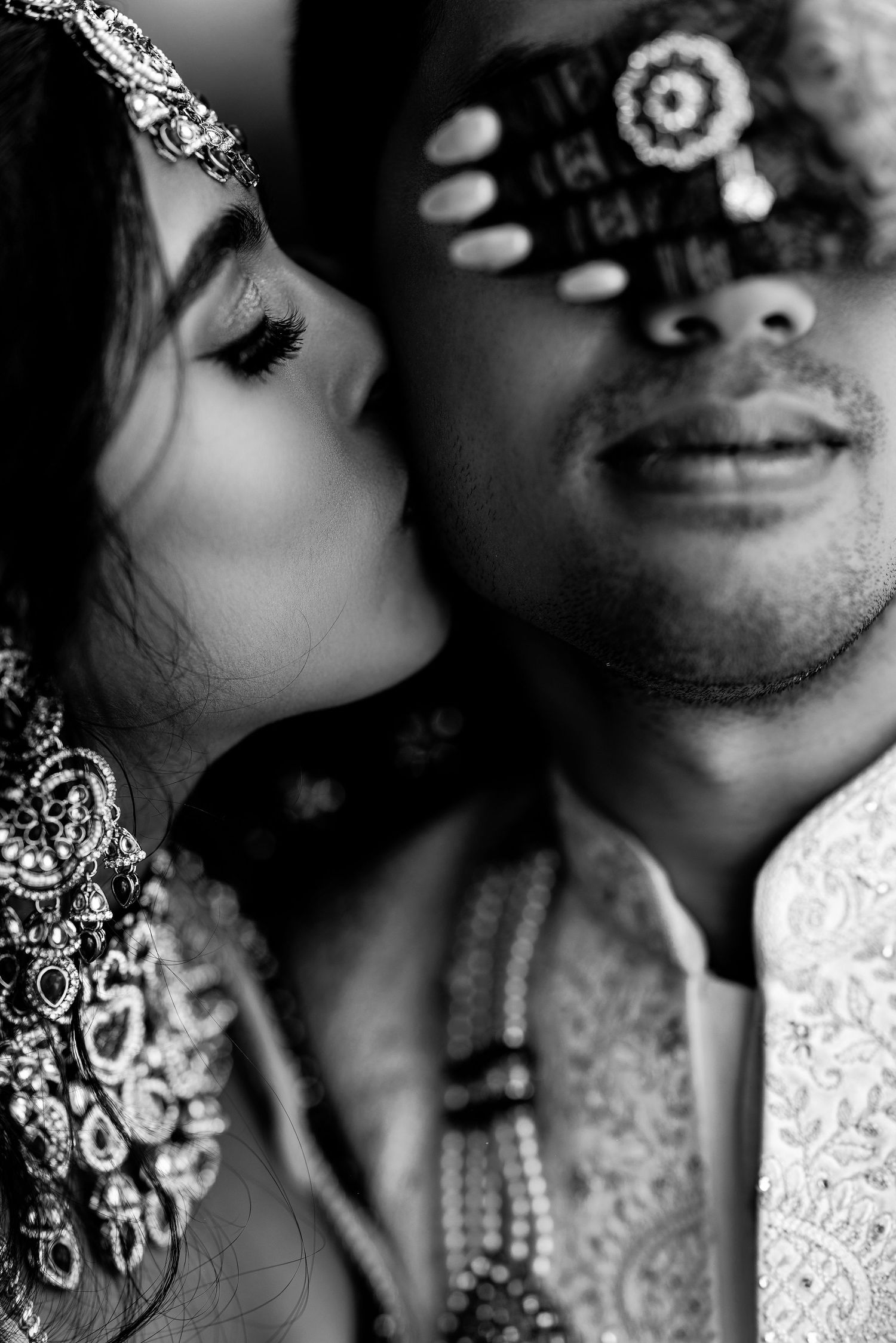 Black and white photo of Indian bride kissing groom's cheek, both adorned in traditional wedding attire and jewelry.