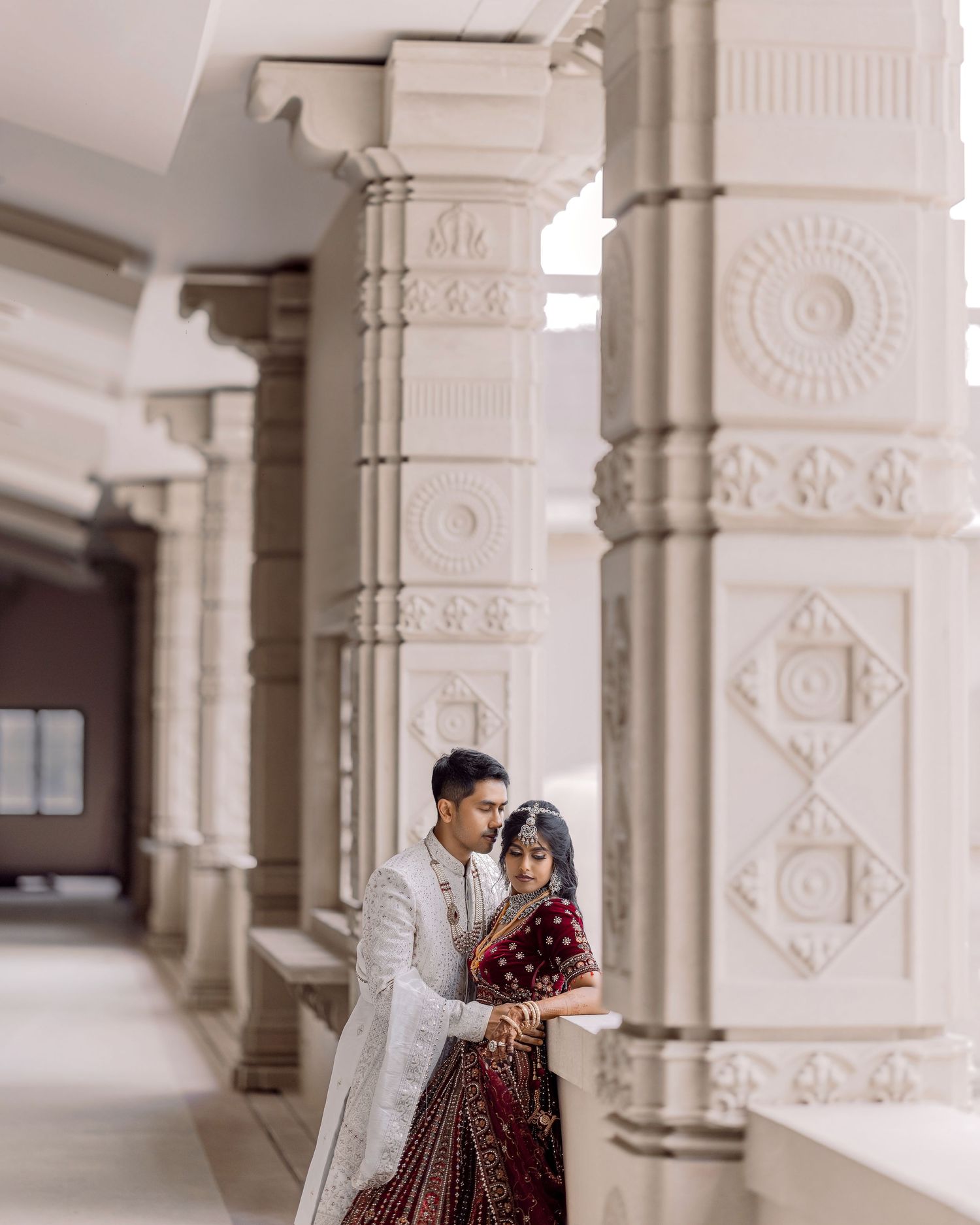 Couple in traditional Indian wedding attire posing between ornate white carved pillars at a Hindu temple.