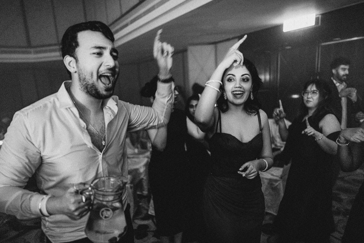 Black and white photo of joyful couple dancing at a party, man holding drink, woman in elegant dress, guests in background.