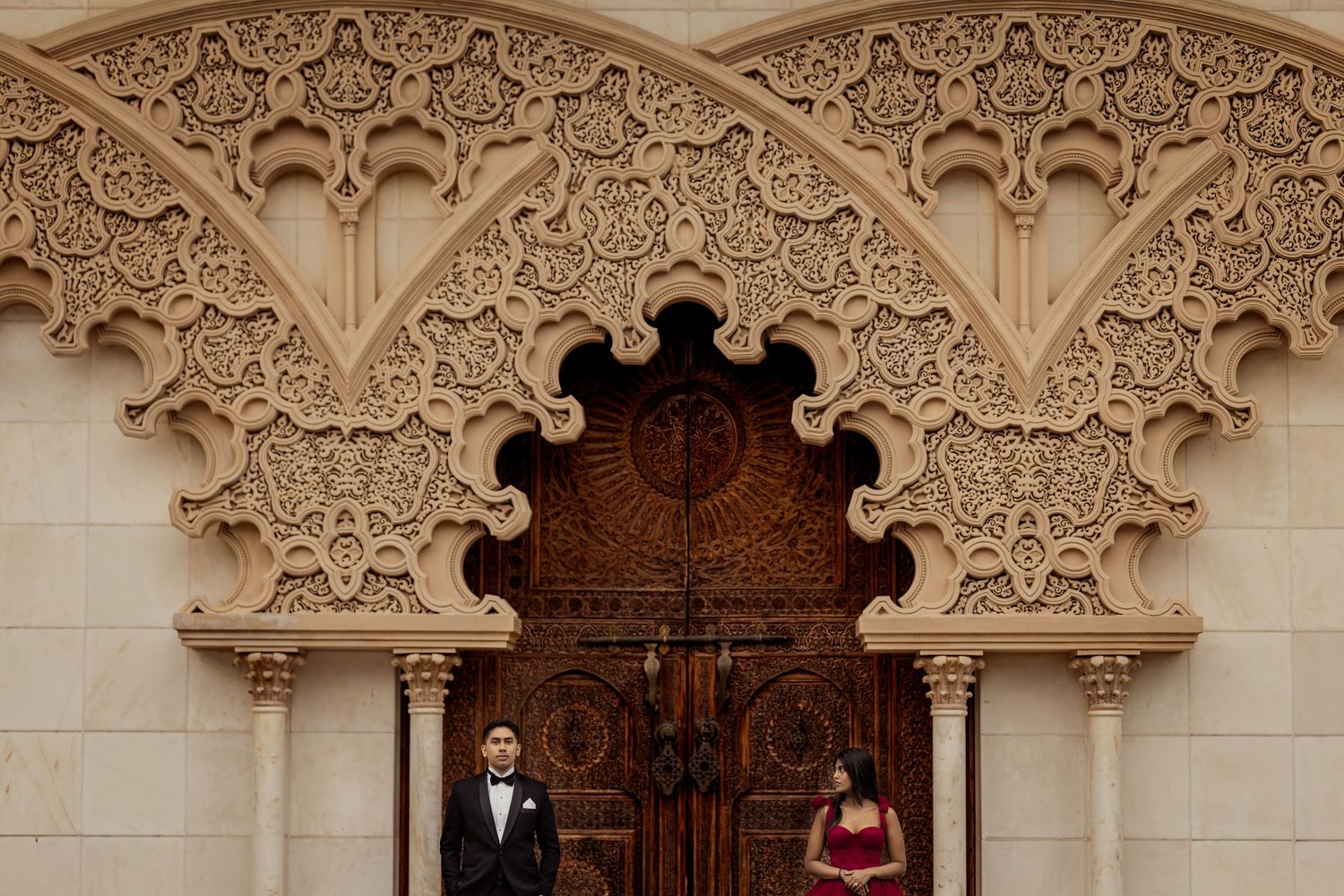 Elegant couple stands beneath ornate Moorish-style archway with detailed carved stonework and wooden door.
