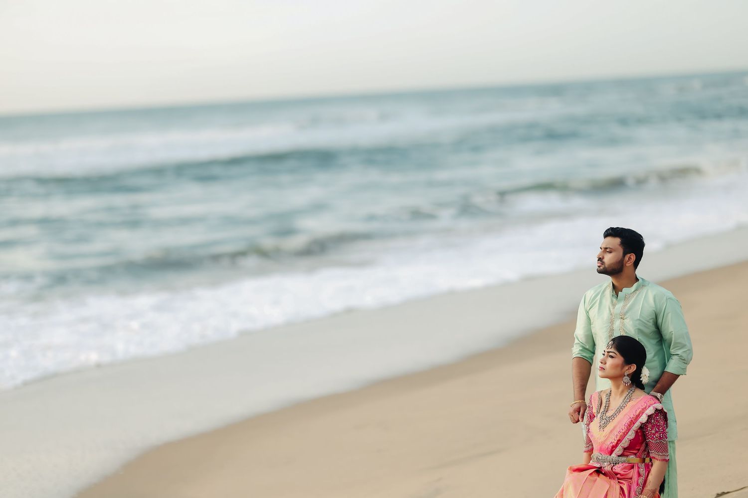 A couple strolls along a serene beach with waves crashing in the background during golden hour.